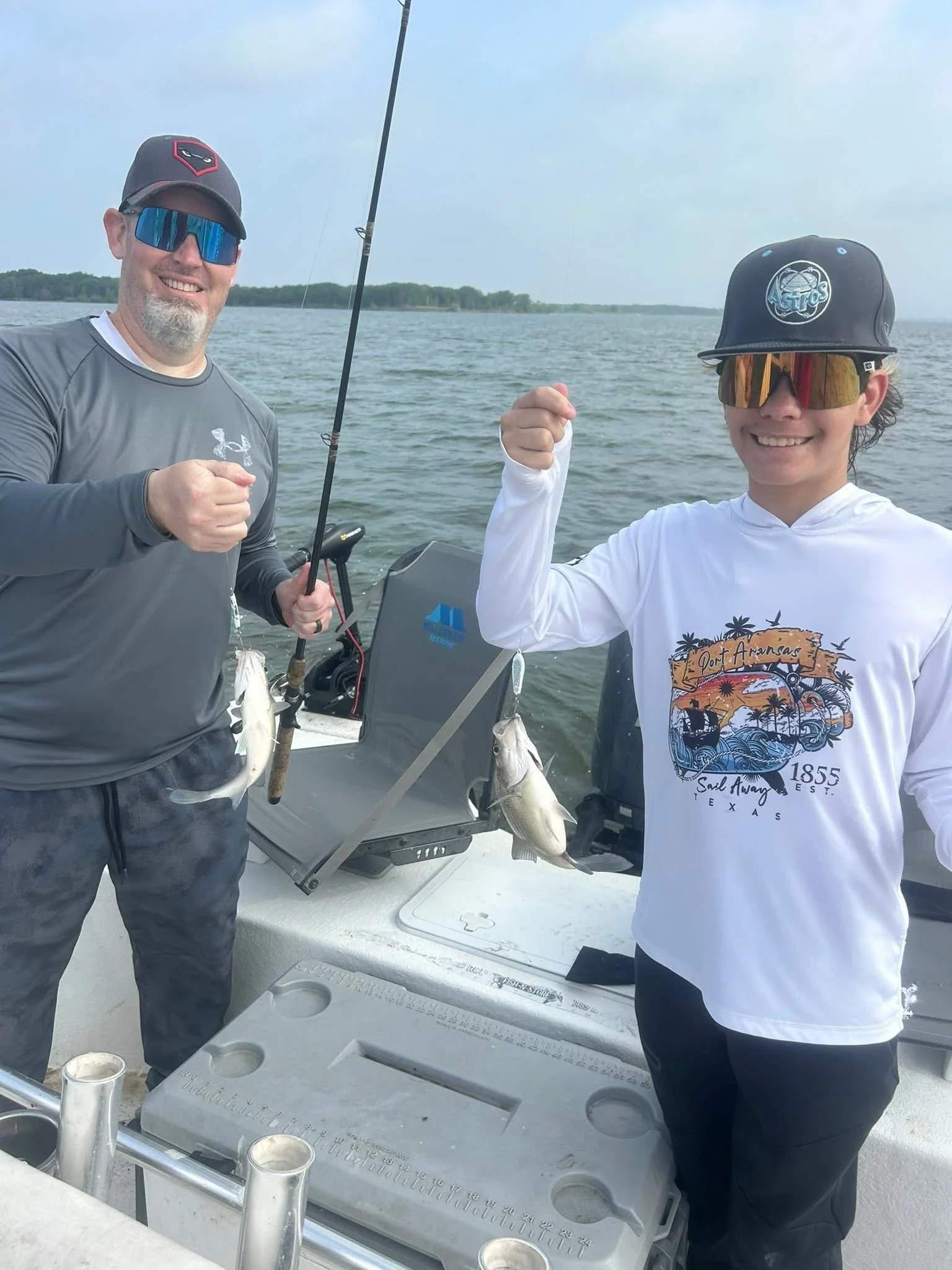 Two men on a boat holding up fish they caught, with fishing gear and water in the background.