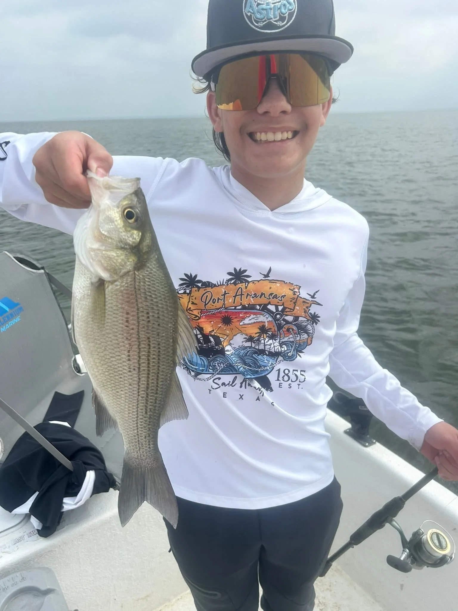 Smiling person wearing a cap with the word 'Astro' and polarized sunglasses, holding a fish they caught, standing near water with cloudy sky in the background.