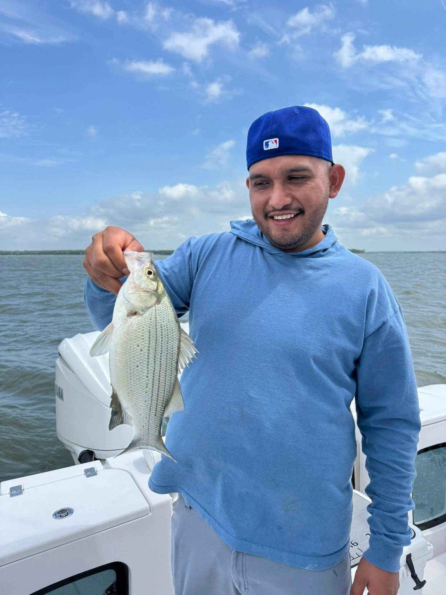 A man wearing a blue hoodie and a blue baseball cap is showing a fish he caught, aboard a boat on a body of water with a cloudy sky in the background.