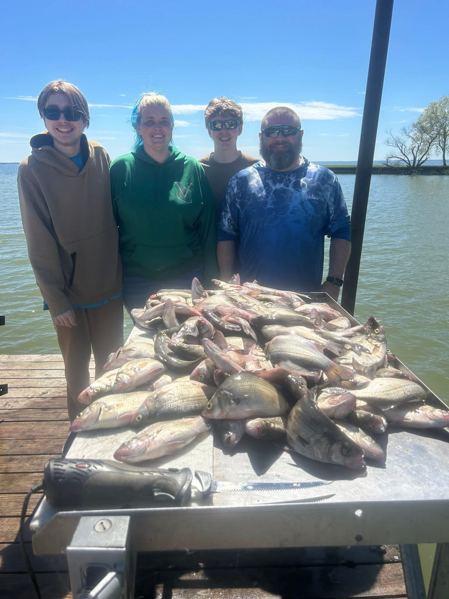 Four people standing on a dock by the water with an array of freshly caught fish on a table in front of them, smiling for the photo on a sunny day.
