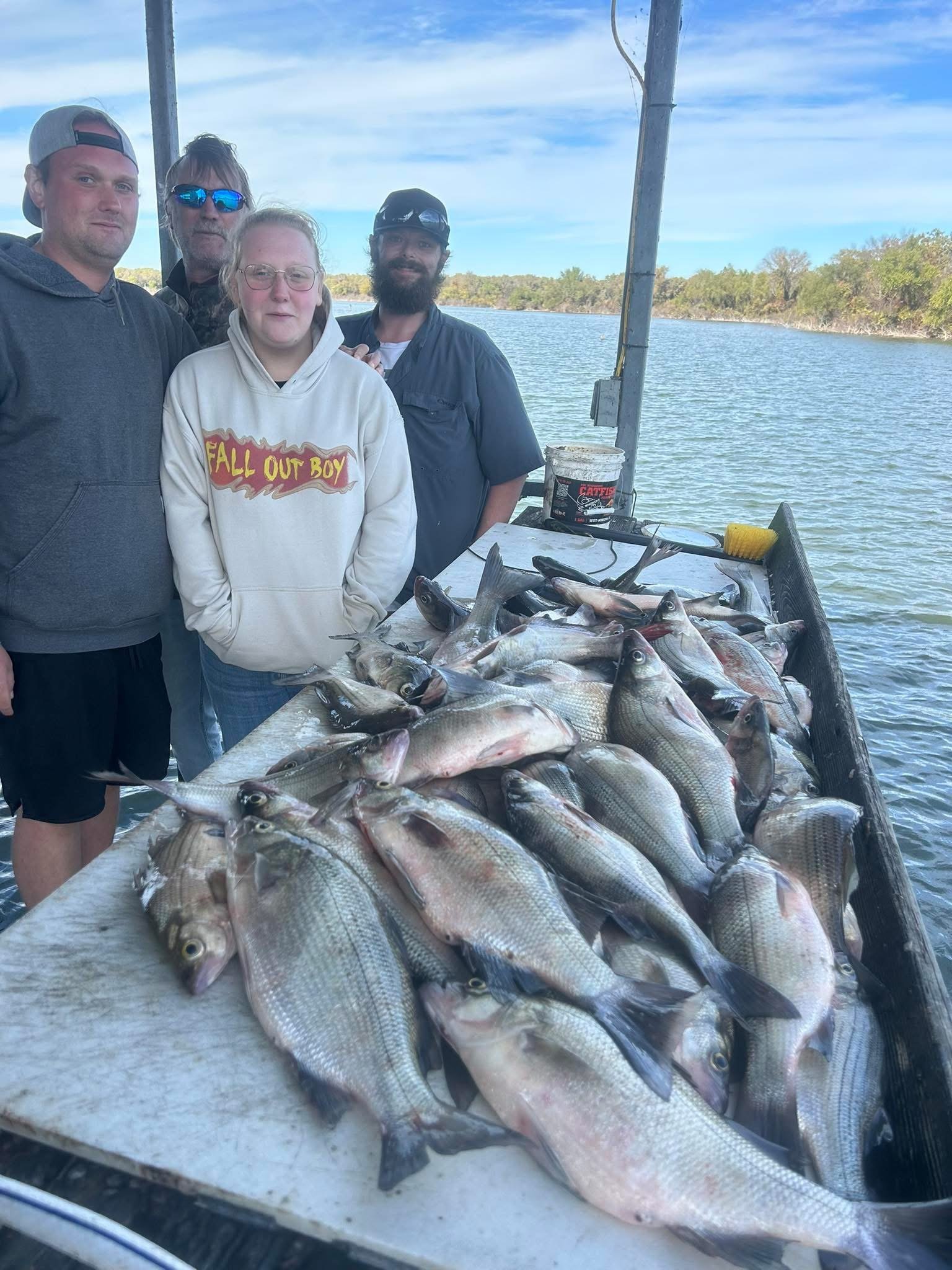 Four people standing behind a large pile of fish on a boat, with water and trees in the background.