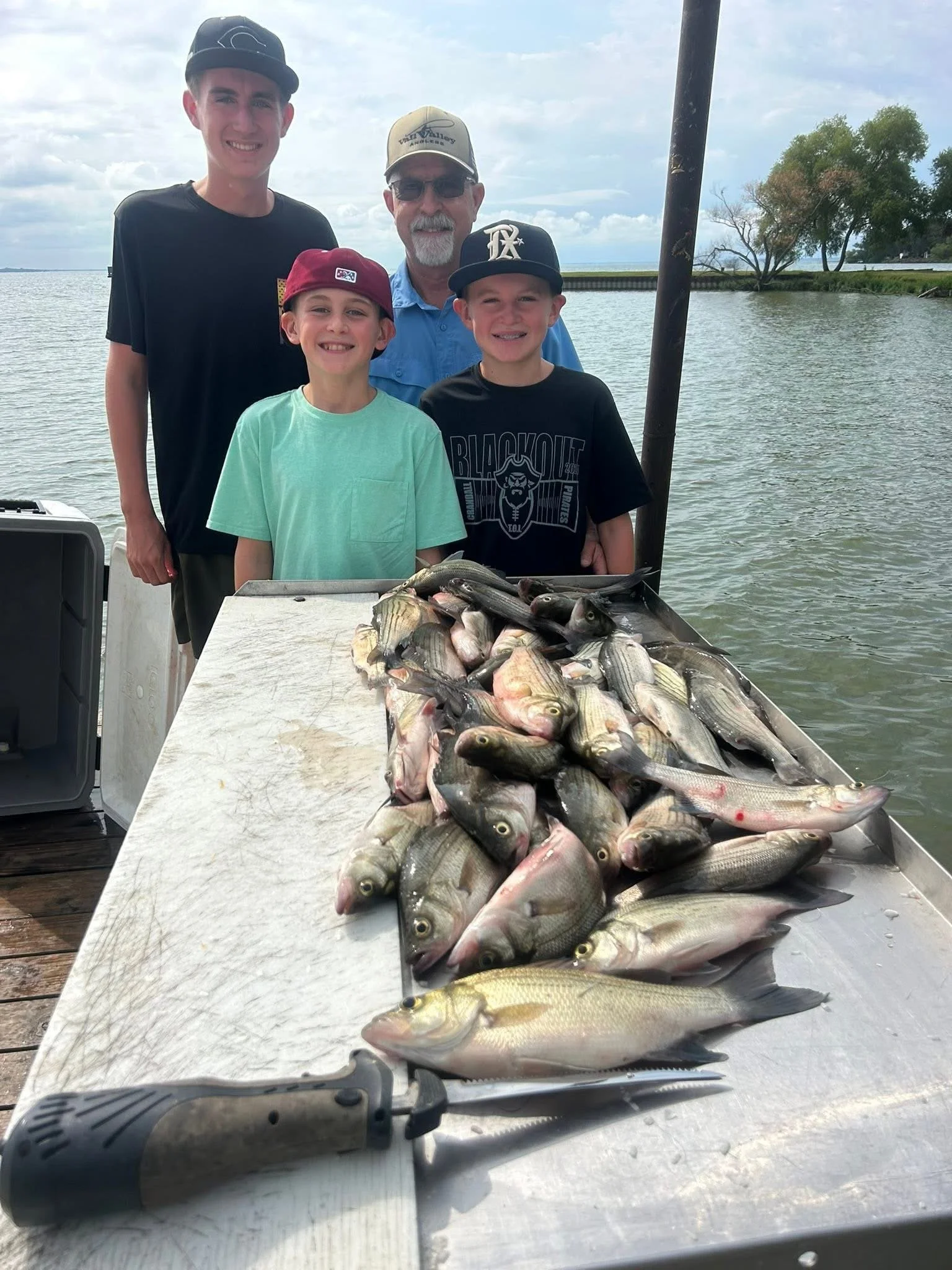 Four males, two children and two adults, standing outdoors by water, displaying caught fish on a table, with a cloudy sky and trees in the background.