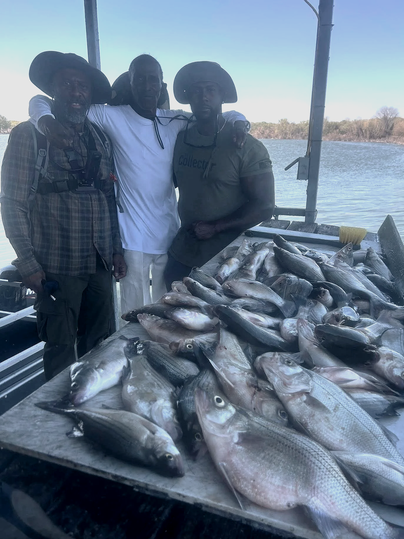 Three men standing behind a table full of freshly caught fish outdoors near a body of water with trees in the background.
