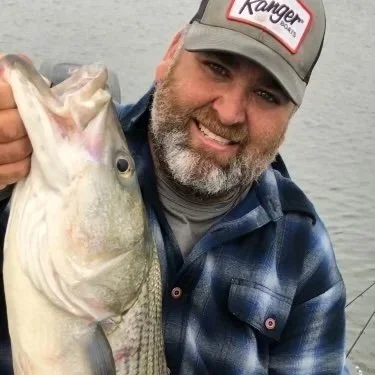 A man smiling while holding a large fish near a body of water.
