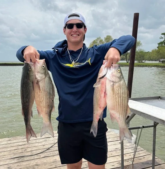 Man in sunglasses and a navy jacket holding two large fish, one in each hand, standing on a dock by the water on an overcast day.