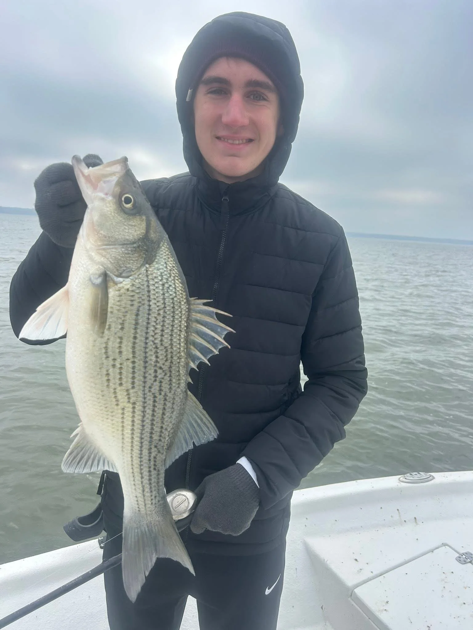Young man in black jacket and gloves holding a fish on a boat in a large body of water under cloudy sky.