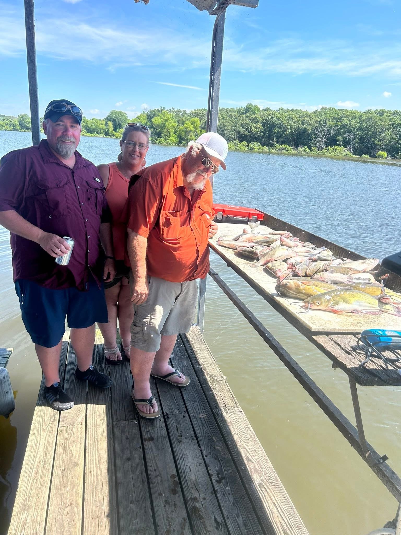 Three people standing on a dock beside a table full of fish, with water and trees in the background, during sunny weather.