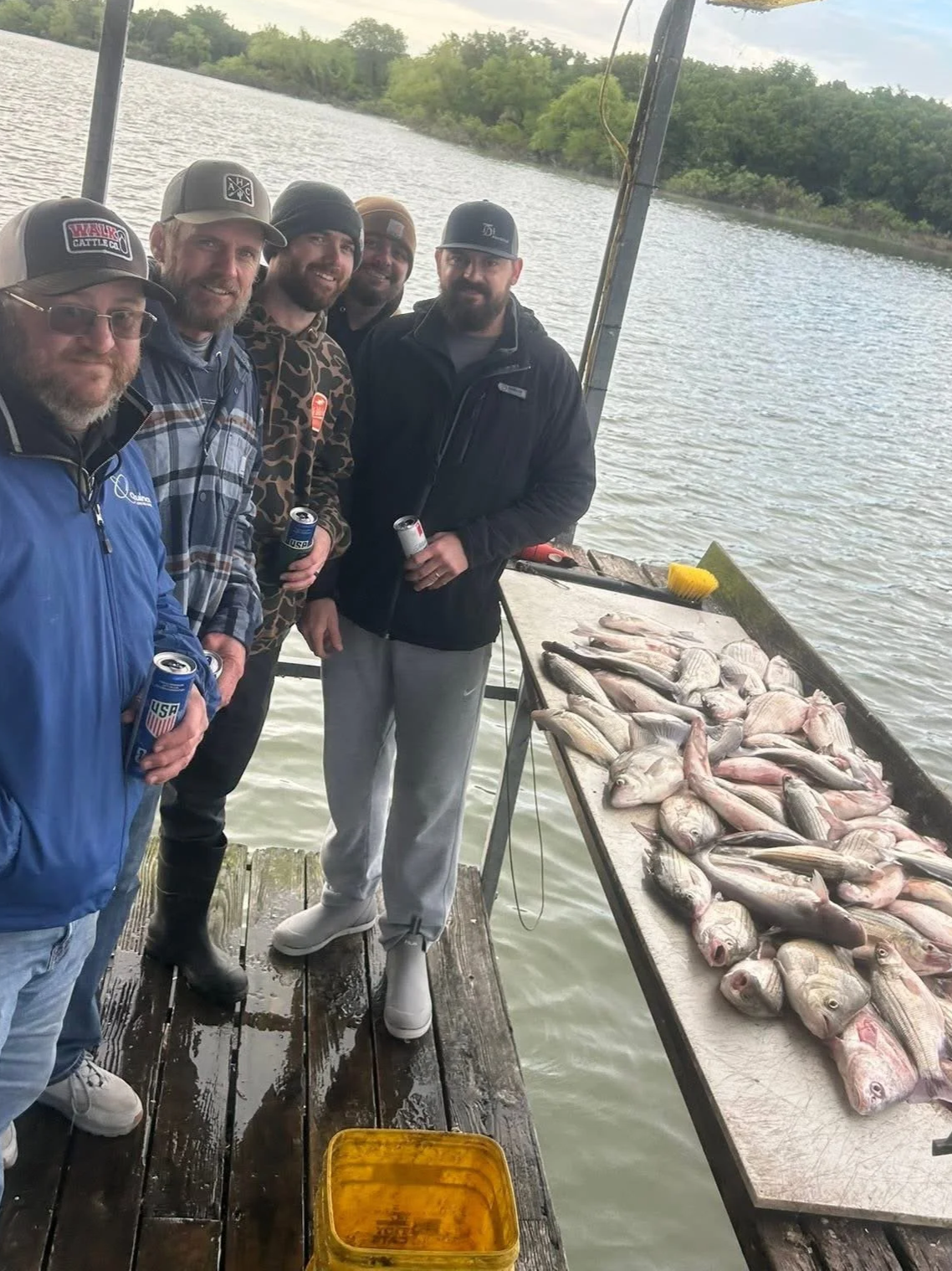 A group of six men standing on a wooden dock beside a table filled with a large catch of fish, holding cans of beer and posing for a photo near a body of water with trees in the background.