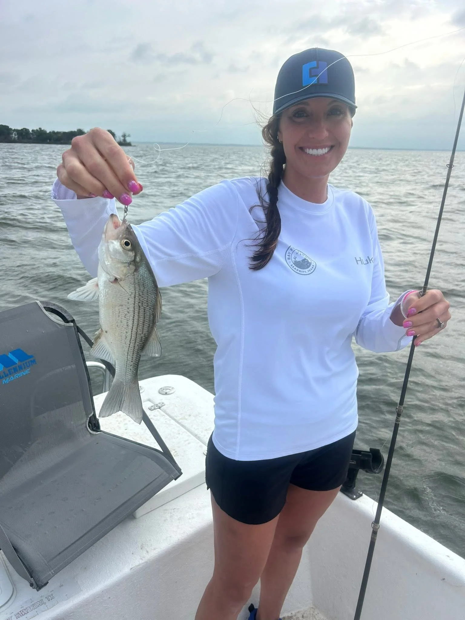 Woman on a boat holding a fish she caught, smiling, wearing a black baseball cap and a white long-sleeve shirt with logos, holding a fishing rod, and the water and sky are visible in the background.