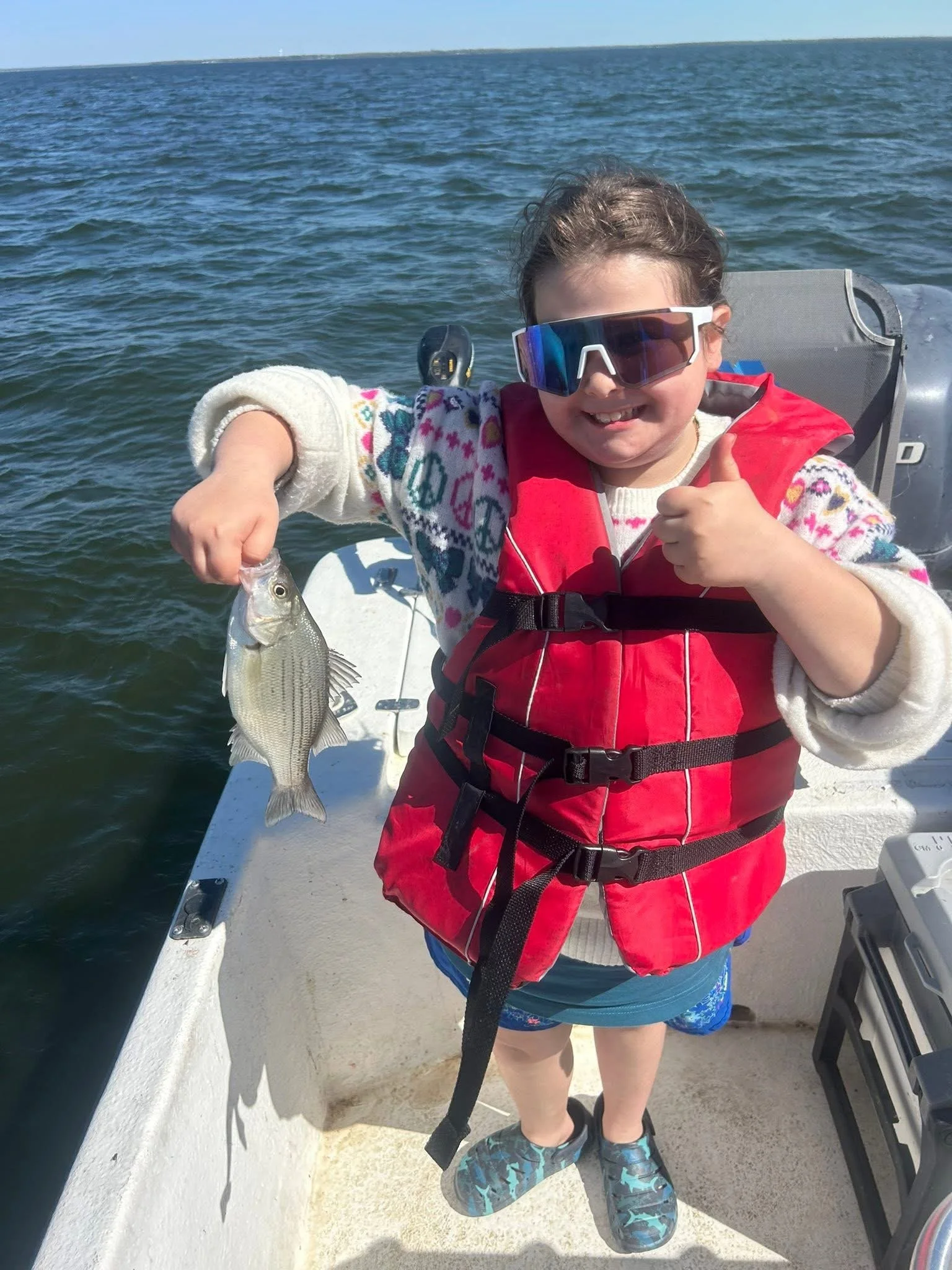A young girl wearing sunglasses and a red life jacket, smiling and holding a fish, on a boat in the water.