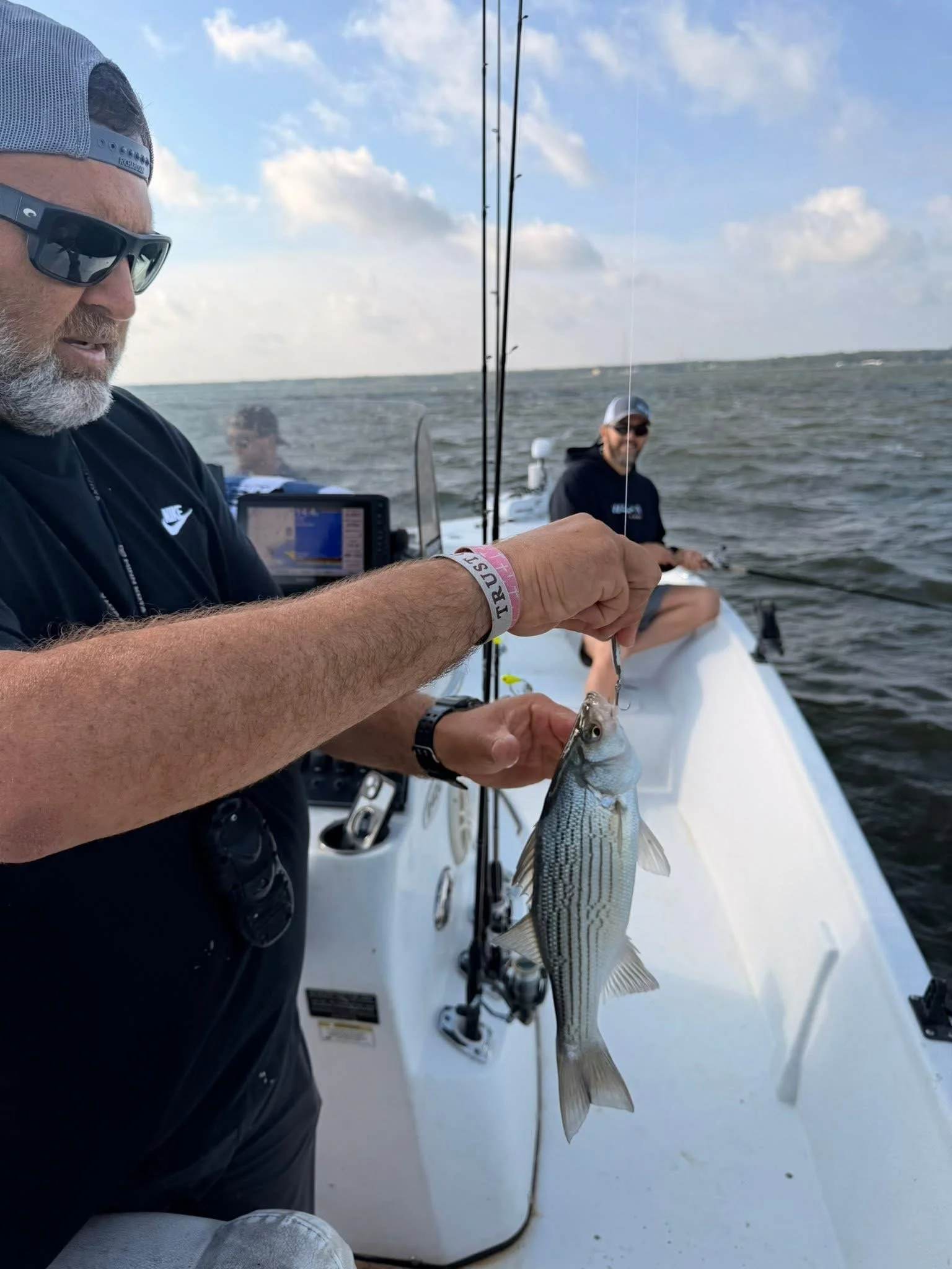 A man with sunglasses and a gray cap holding a fish on a boat during daytime, with another person in the background and open water under a partly cloudy sky.