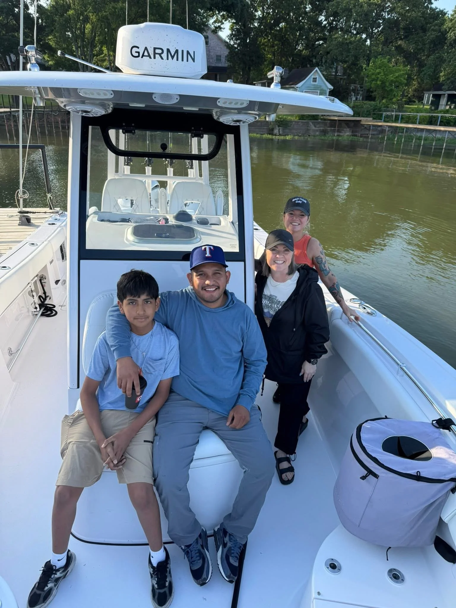 Four people smiling on a boat, with trees and houses visible in the background.