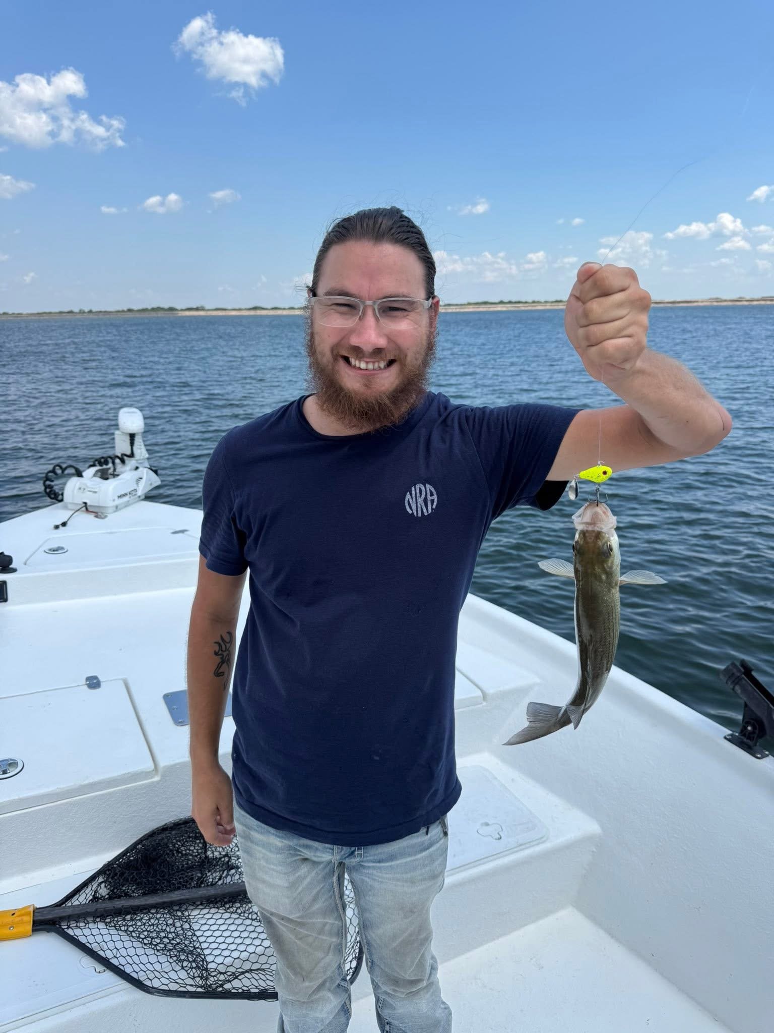 Man with glasses and a beard smiling on a boat holding up a fishing line with a small fish caught on the hook. The water and sky are visible in the background.
