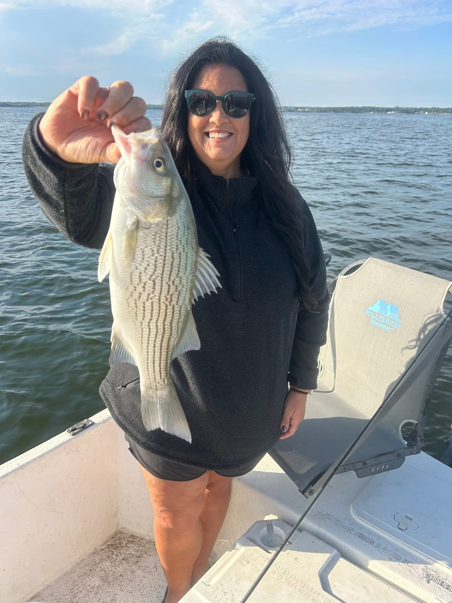 Woman with long dark hair, wearing sunglasses, holding a fish she caught on a boat in a body of water, with a distant shoreline and partly cloudy sky in the background.