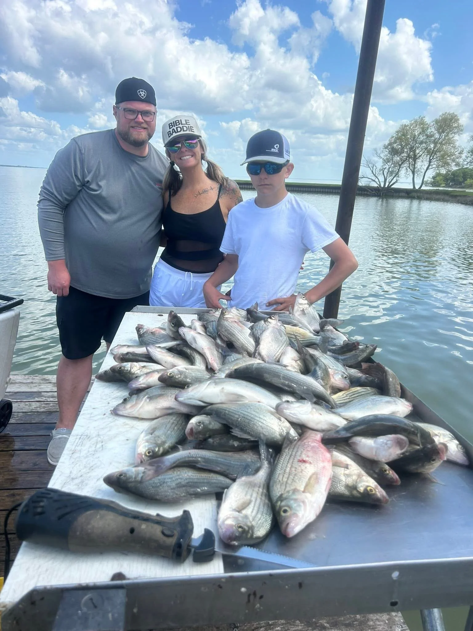 Three people standing behind a large table filled with caught fish on a dock by a body of water, smiling, with blue skies and clouds.