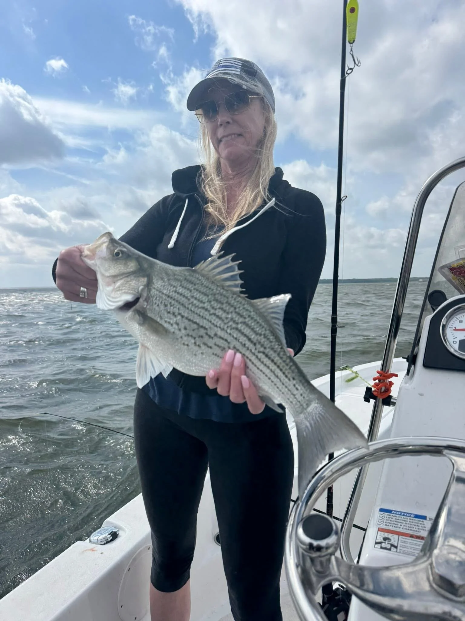 A woman wearing sunglasses, a cap, and a black hoodie is on a boat holding a fish she caught. The sky is partly cloudy.