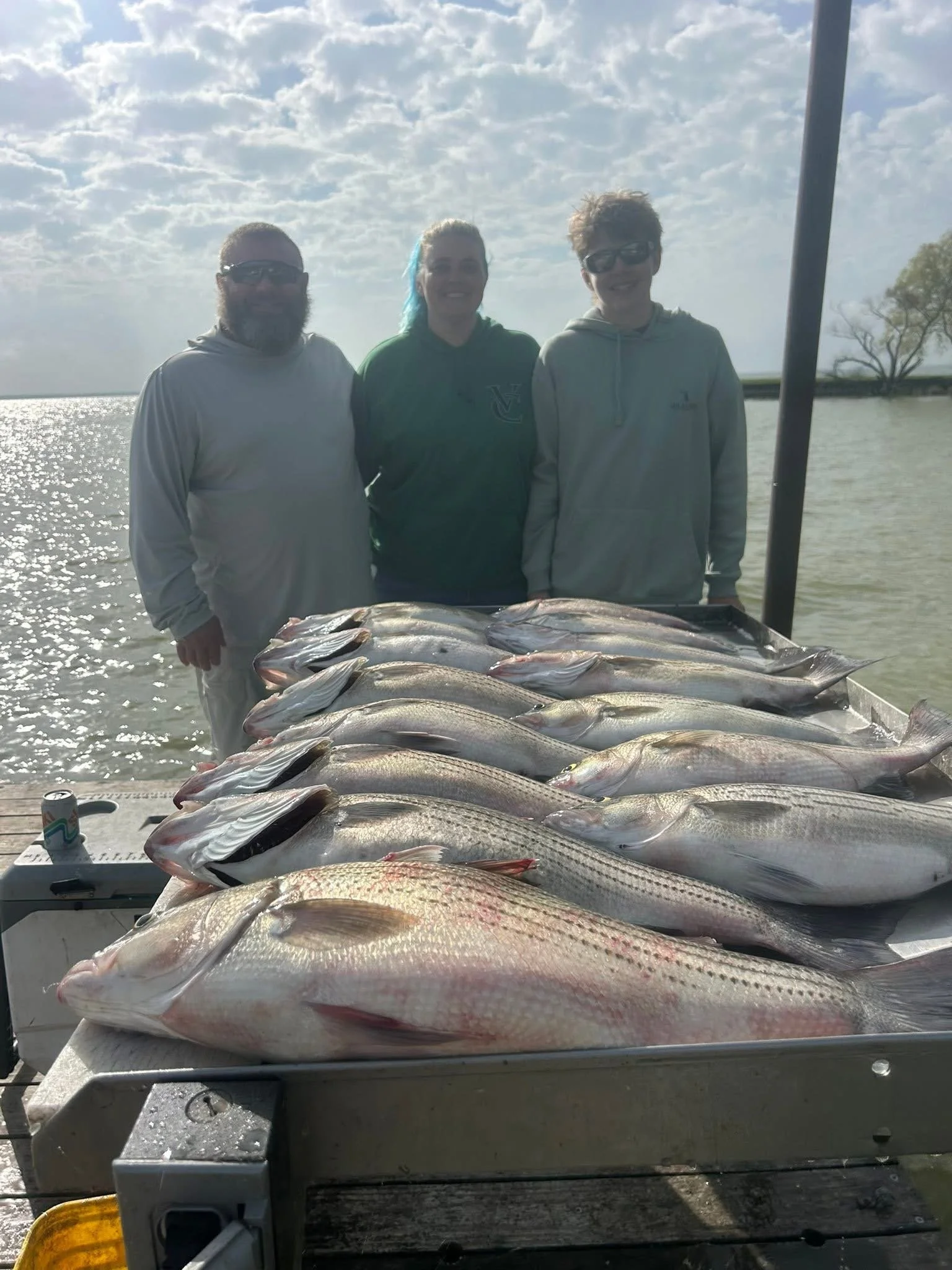 Three people standing behind a table full of fish, outdoors near a body of water with cloudy skies.