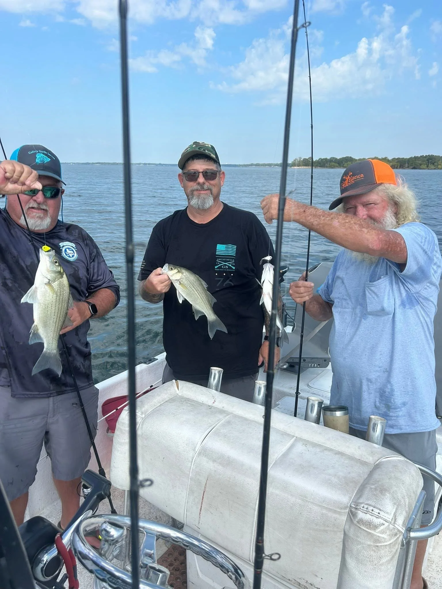 Three men on a boat holding fish they caught while fishing during the day. The water and sky are visible in the background.
