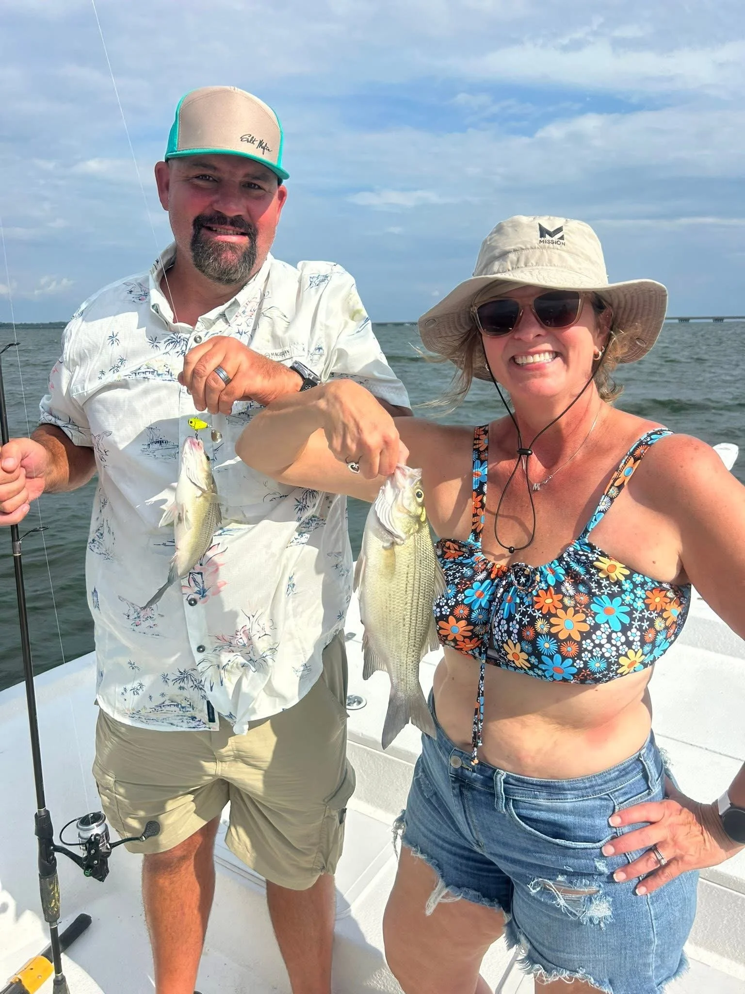 A man and woman on a boat holding fishing rods and showing off two fish they caught. The woman is wearing a sun hat and sunglasses, and the man is wearing a cap. The background shows a body of water and a distant bridge under a partly cloudy sky.