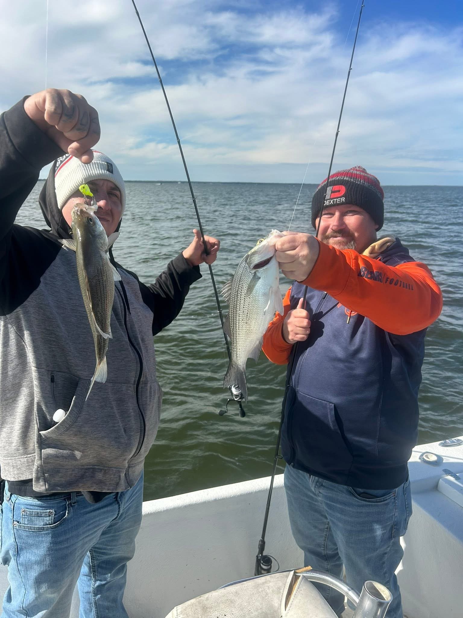 Two men on a boat holding up their fishing catches. One man is holding a small fish with a fishing lure, and the other man is holding a larger fish. They are dressed warmly and wearing beanies, with a body of water and a cloudy sky in the background.