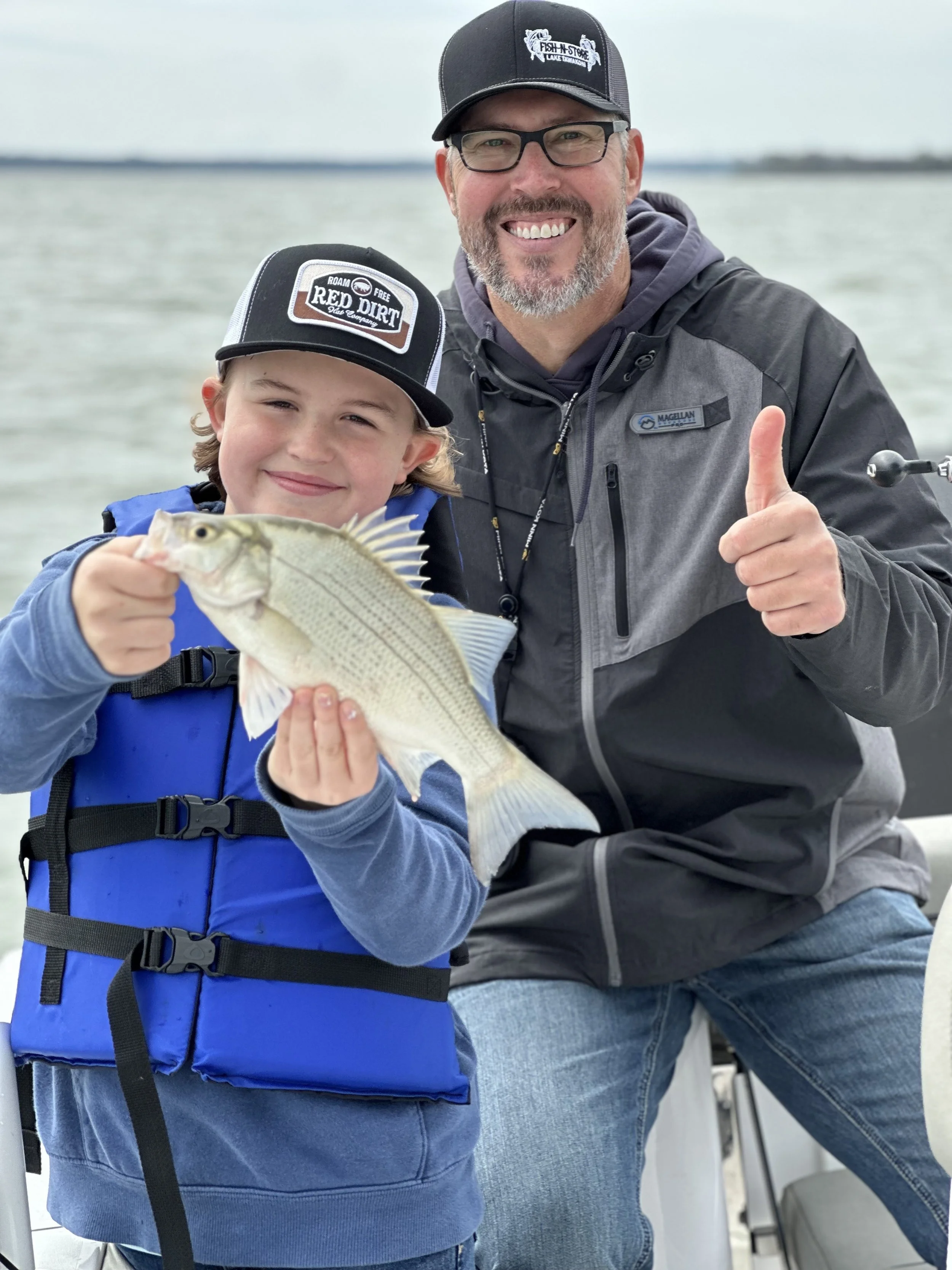A smiling man with glasses, wearing a black cap and jacket, giving a thumbs-up while on a boat or dock, standing close to a young boy with a cap and blue life jacket, holding a fish in his hand.
