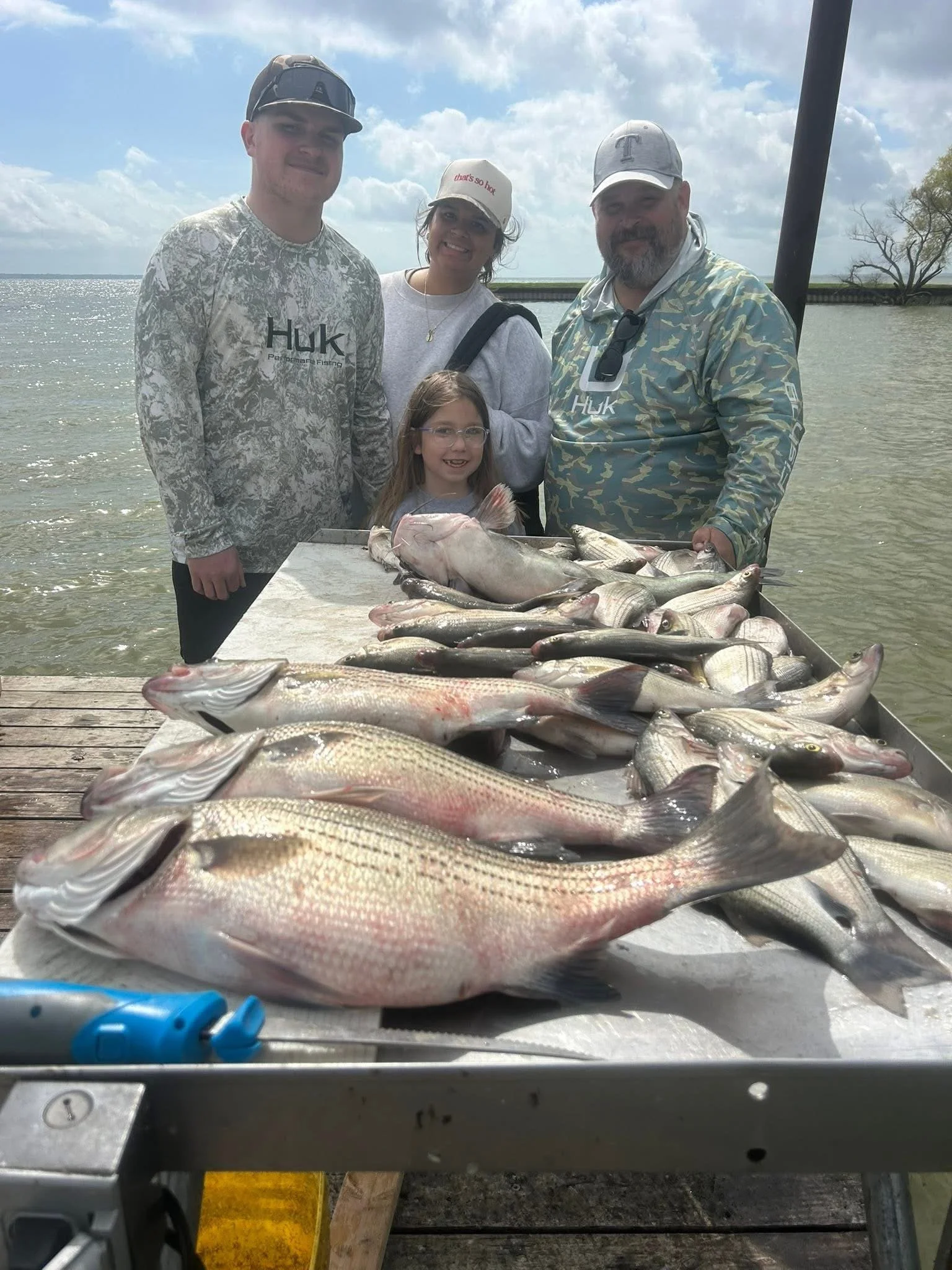 A group of four people, two men, one woman, and a girl, standing behind a table filled with fish outdoors near water, with a cloudy sky and trees in the background.