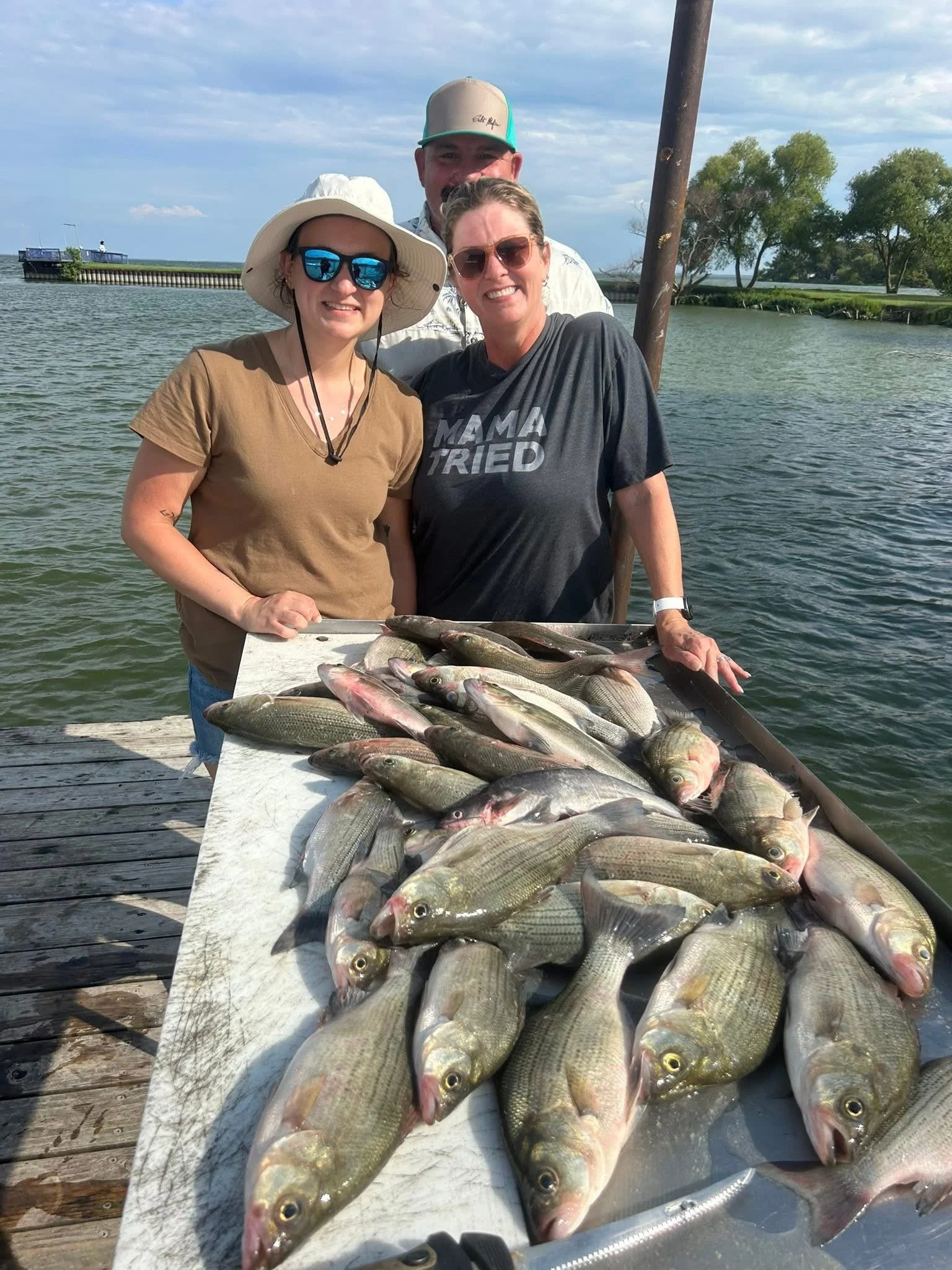 Group of four people on a boat with fishing gear, standing behind a table full of numerous freshly caught fish, with water, trees, and a partly cloudy sky in the background.