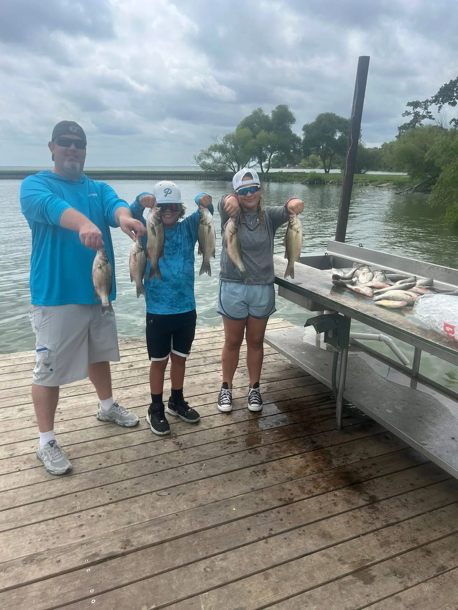 Three people standing on a wooden dock holding caught fish, with a body of water, trees, and cloudy sky in the background.