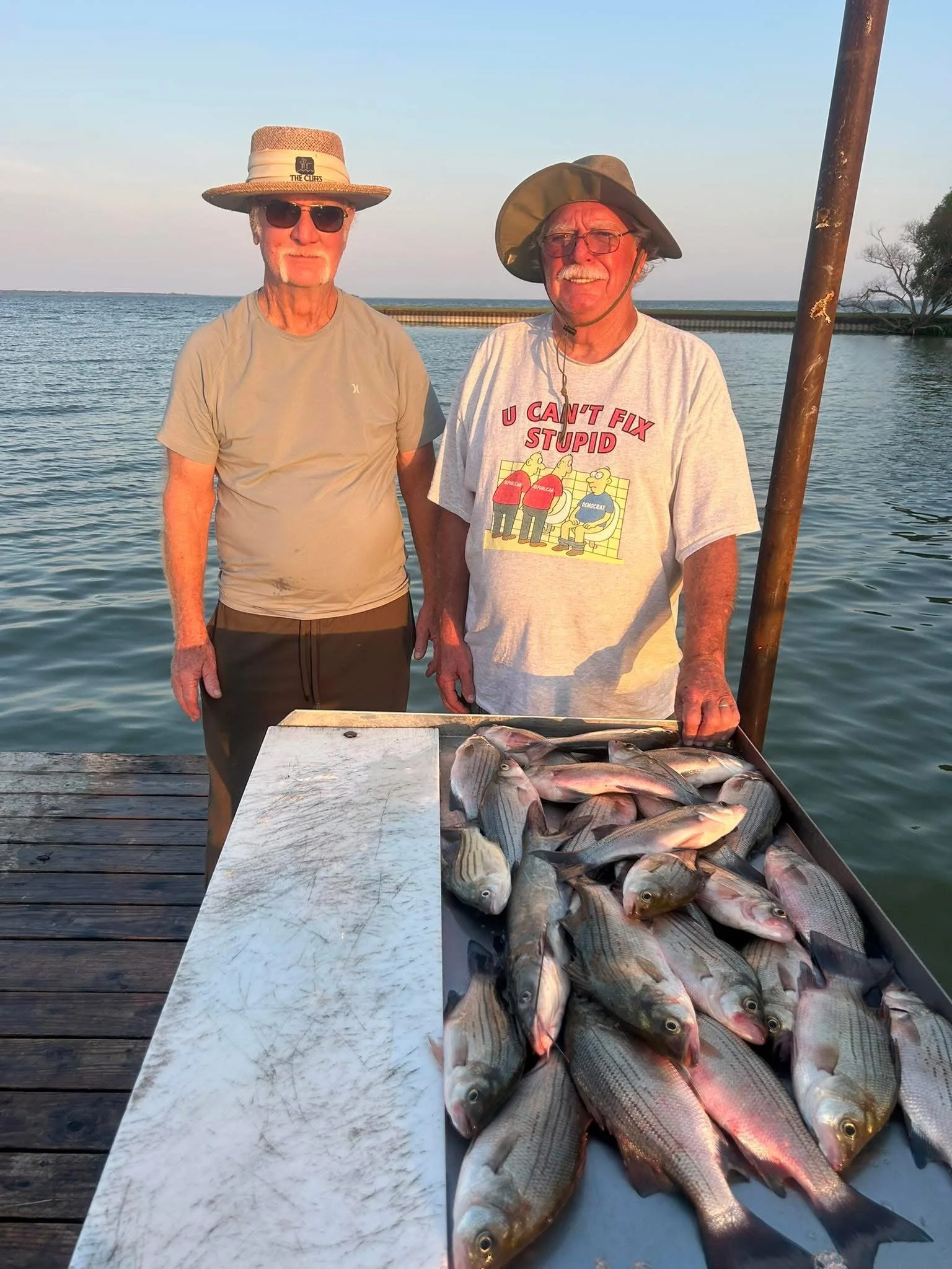 Two elderly men standing on a dock by the water with a large catch of fish on a table in front of them. One man is wearing a beige t-shirt, a straw hat, and sunglasses, while the other man is wearing a white t-shirt with a humorous cartoon and text, and a wide-brimmed hat. A rusty pole is held by the man on the right.