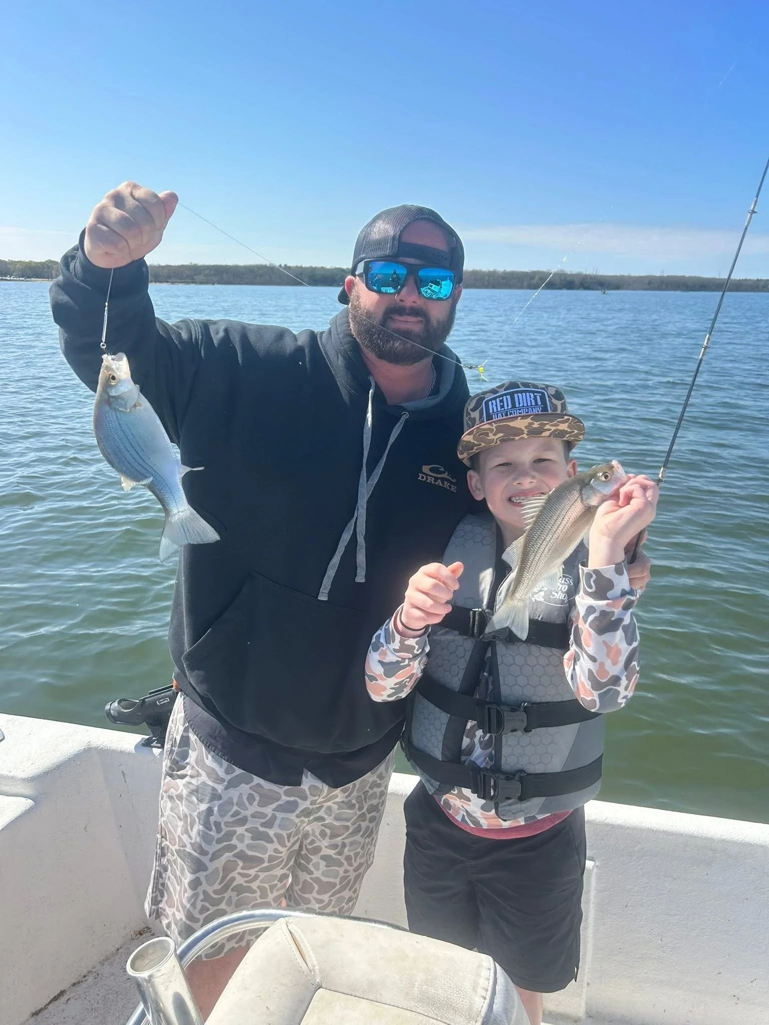 Two people, a man and a boy, stand on a boat holding up two fish they caught, with water and land in the background.