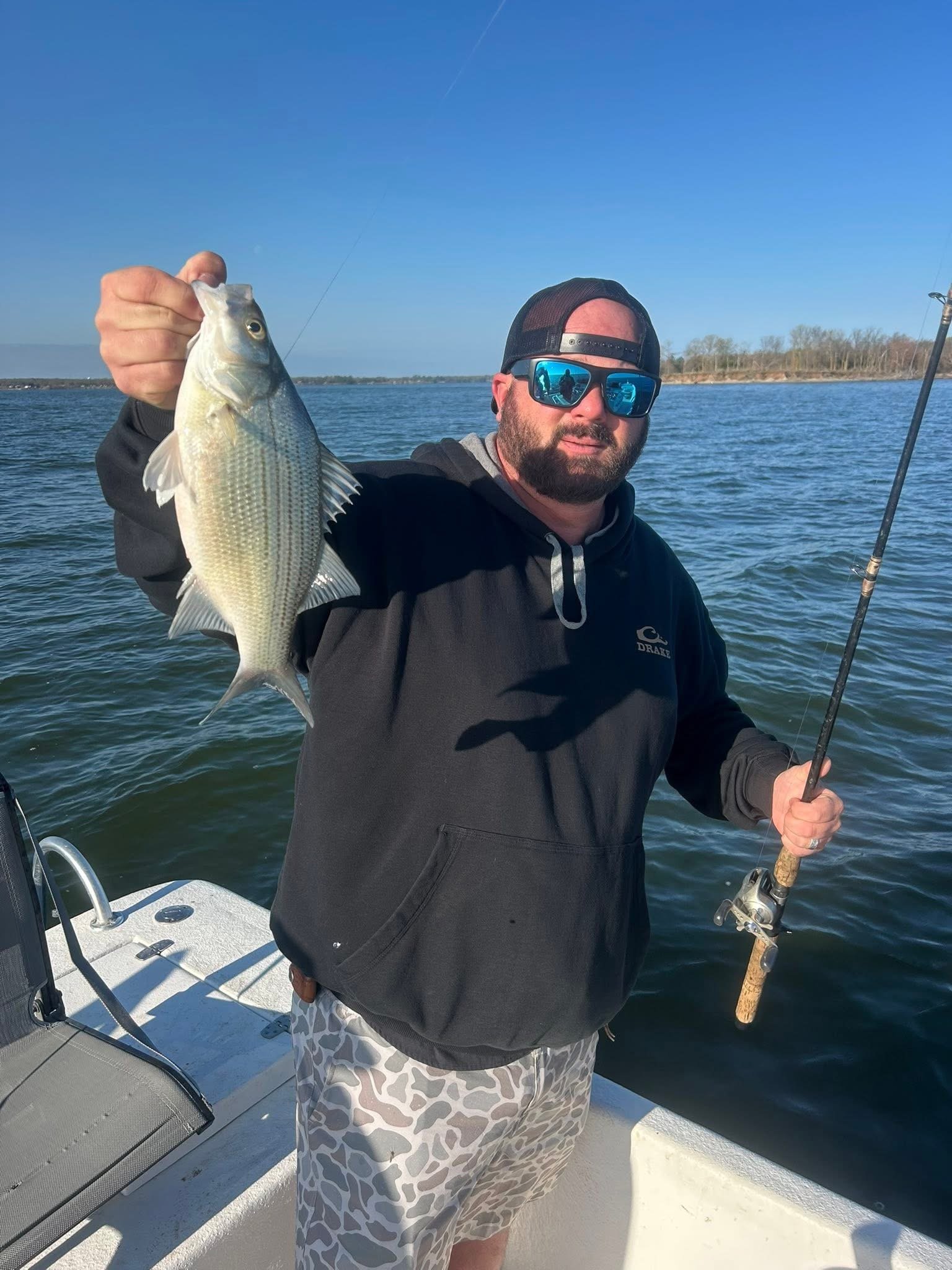 A man in a black hoodie, sunglasses, and a backwards cap is holding a fish on a boat. He is smiling and the water and some land are visible in the background.