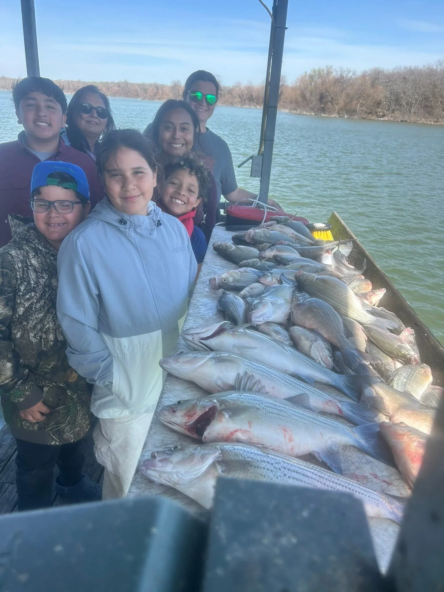 A group of eight people, children and adults, smiling on a boat by a lake during daytime with a catch of fish laid out on a table, trees and water in the background.