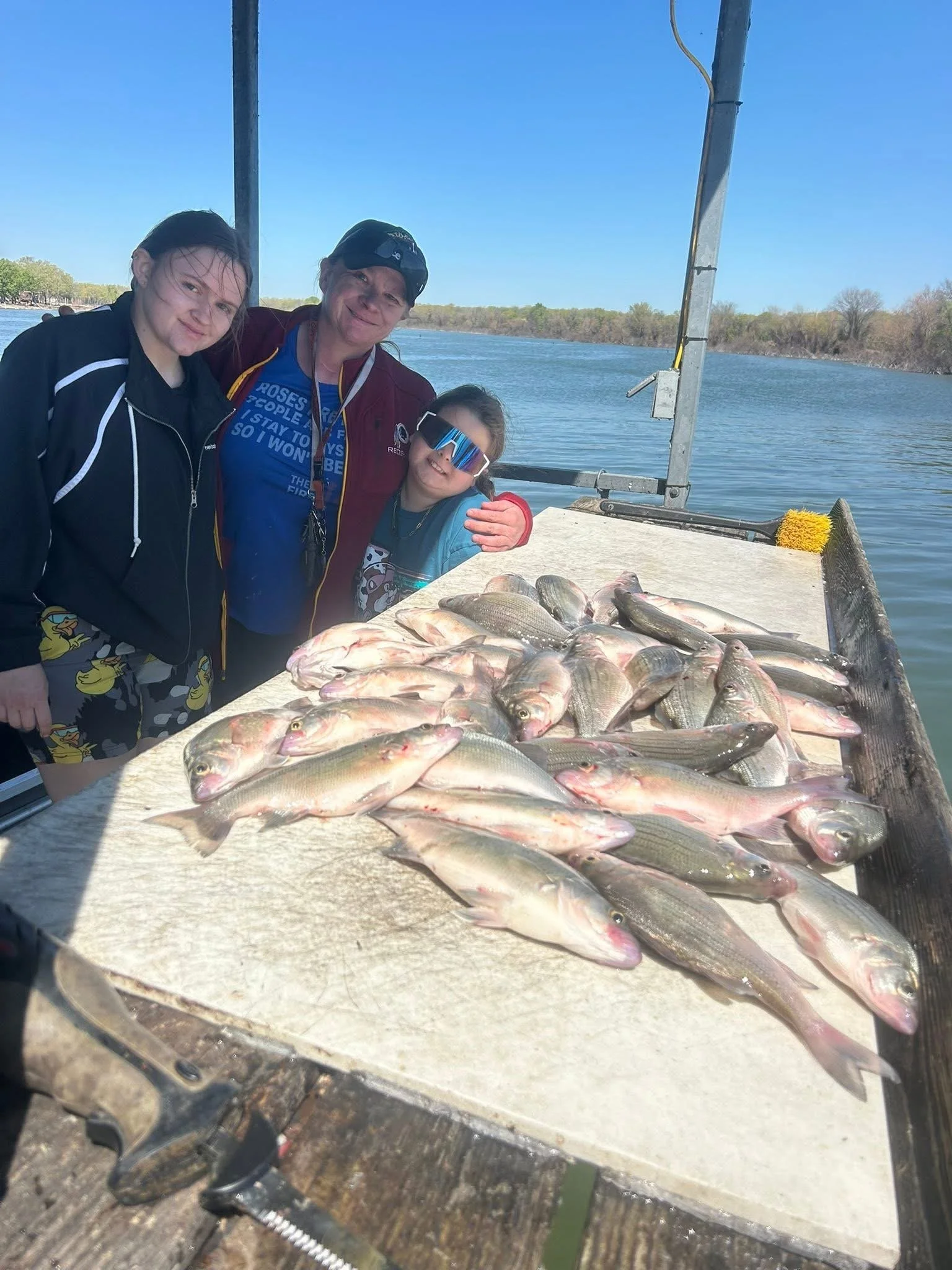 Three people, two young girls and an adult woman, standing behind a large quantity of fish on a dock by a river, with water and trees in the background.