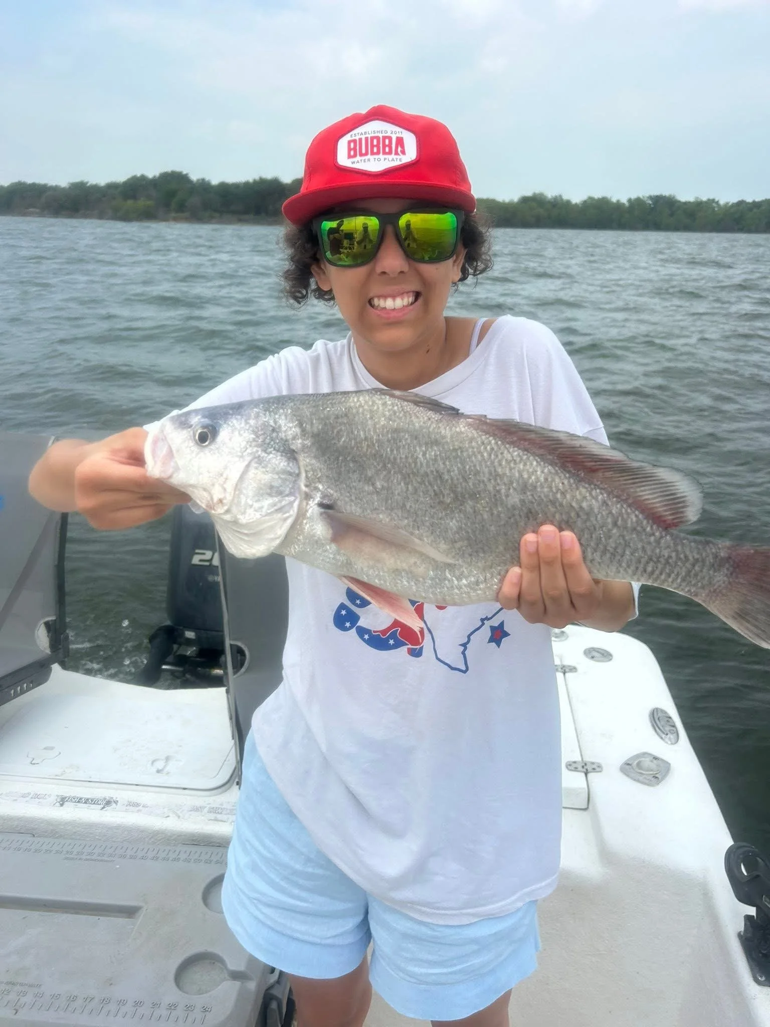 Person on a boat holding a large fish, wearing mirrored sunglasses, a red cap, and a white shirt, with water and greenery in the background.
