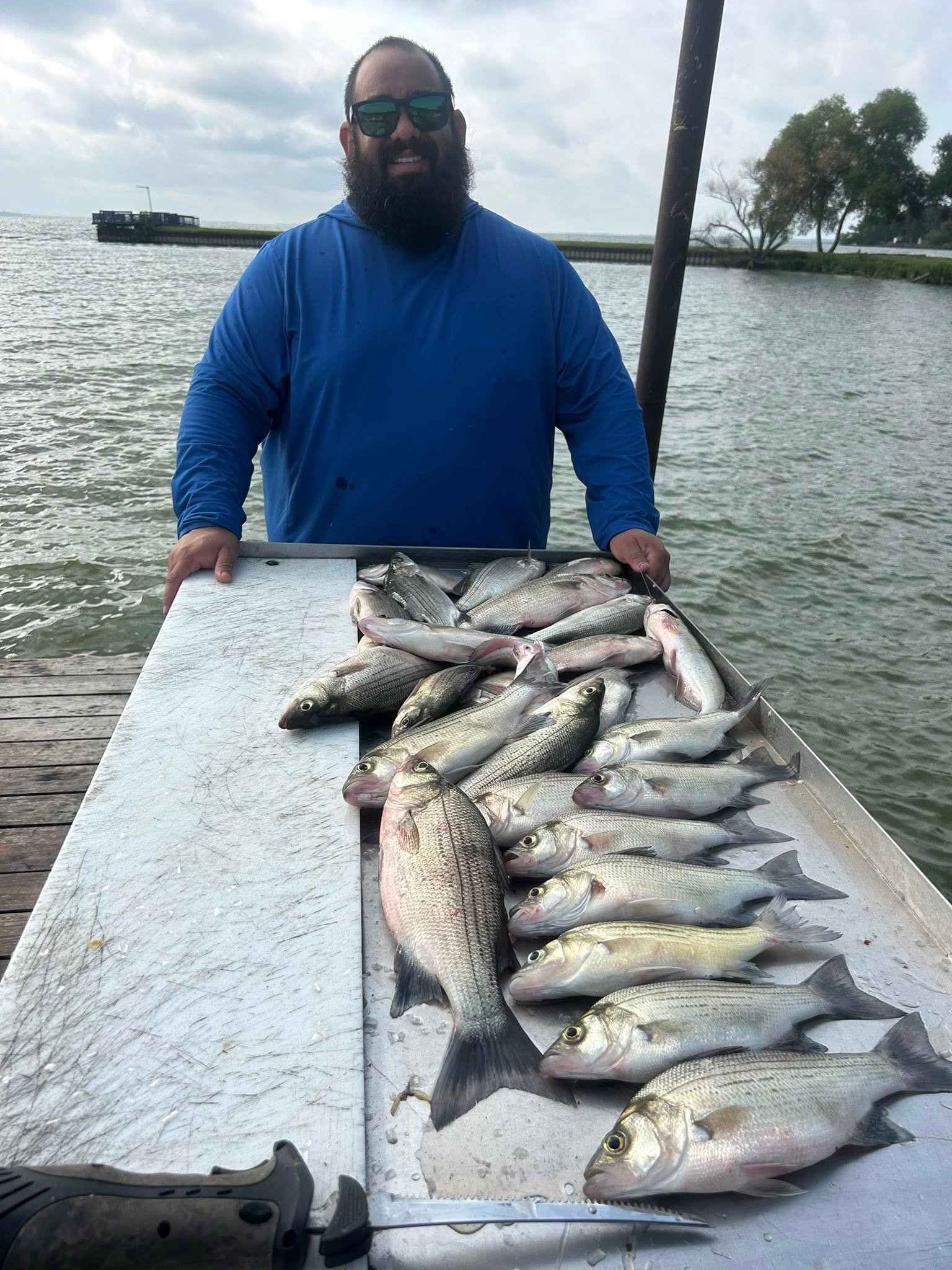 A man with sunglasses and a beard holding a tray of freshly caught fish on a dock by the water.