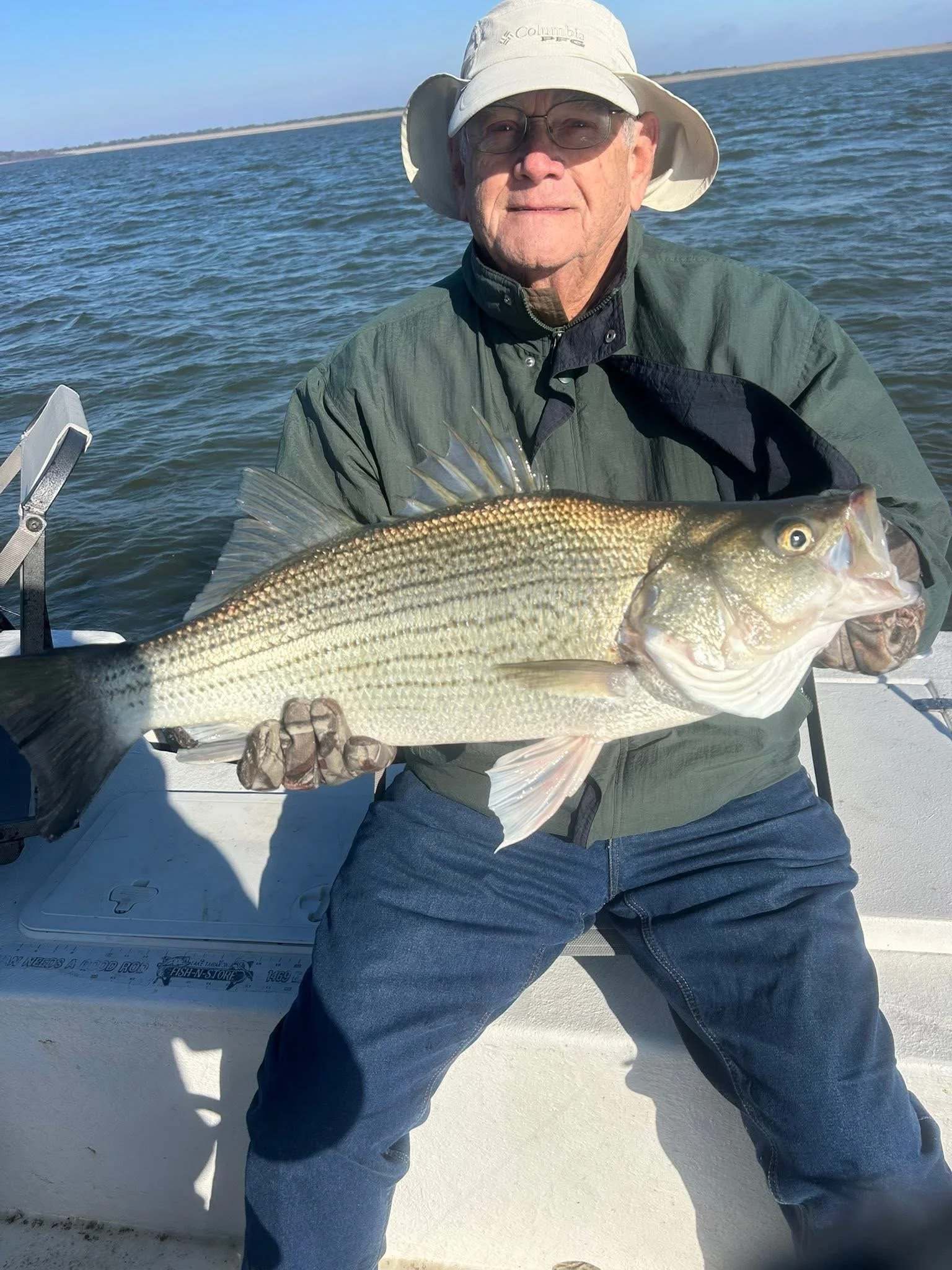 An elderly man wearing a wide-brimmed hat, glasses, and outdoor clothing is sitting on a boat holding a large fish, with water and distant land in the background.