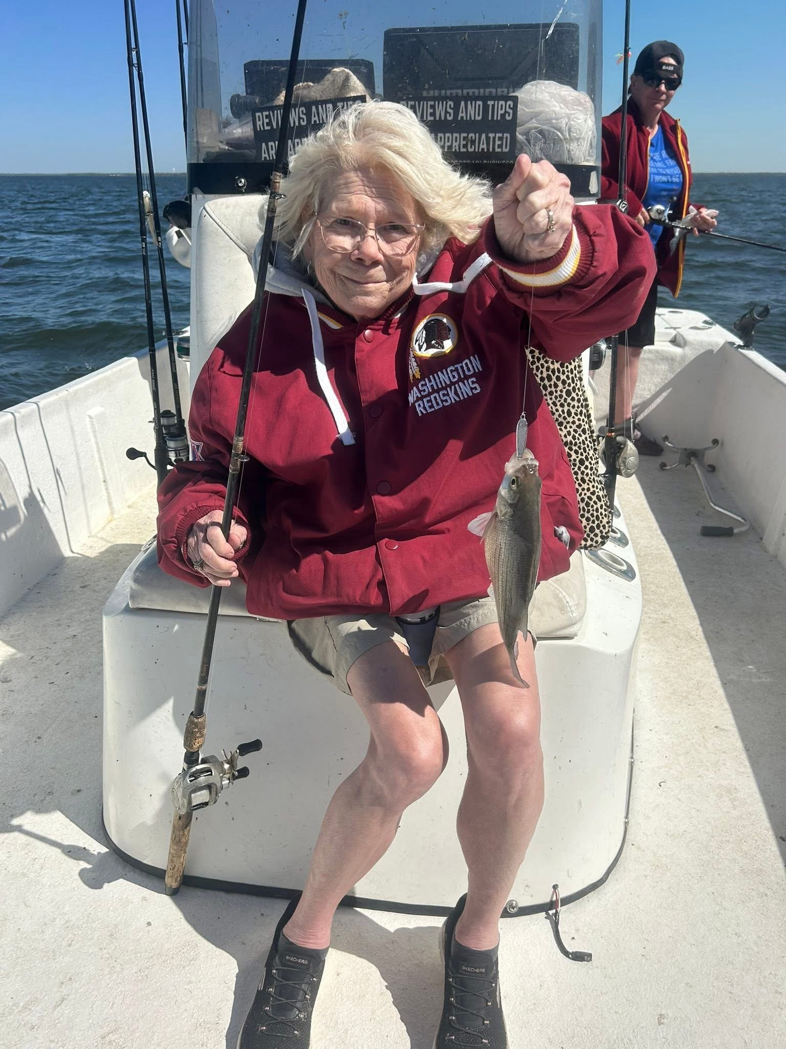 An elderly woman sitting on a boat holding up a fish she caught while fishing. She is wearing glasses and a Washington Redskins jacket. There is a man in the background also fishing.