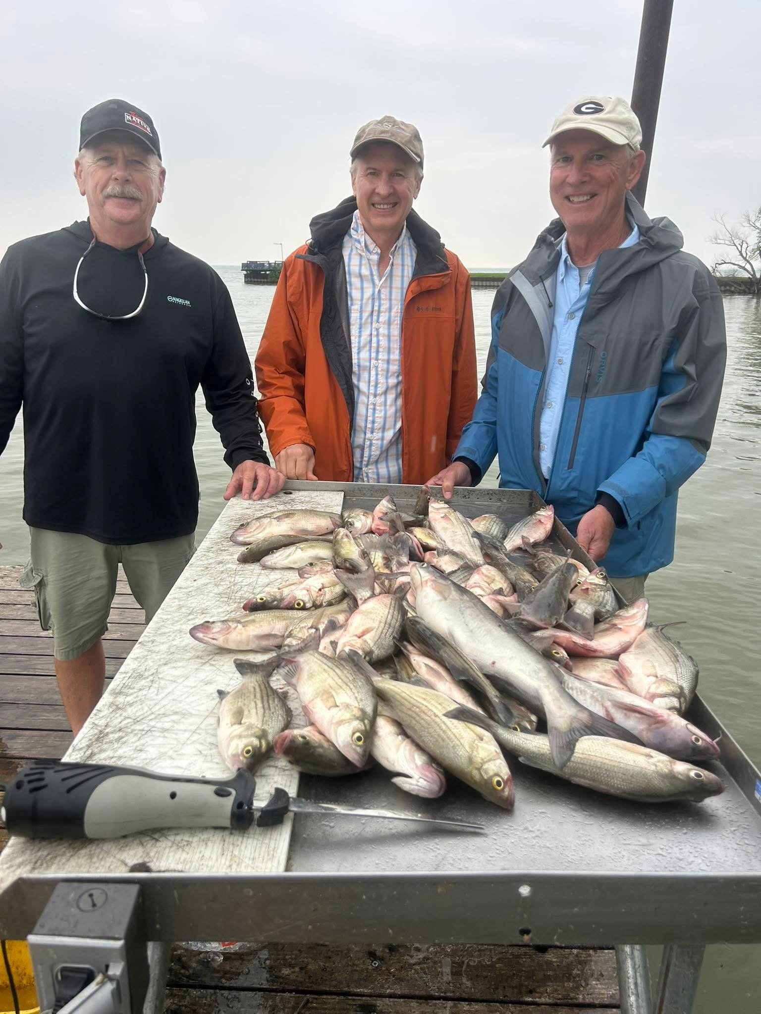 Three men smiling and standing behind a large table filled with freshly caught fish. They are outdoors near water, dressed in jackets and hats, with a cloudy sky overhead.