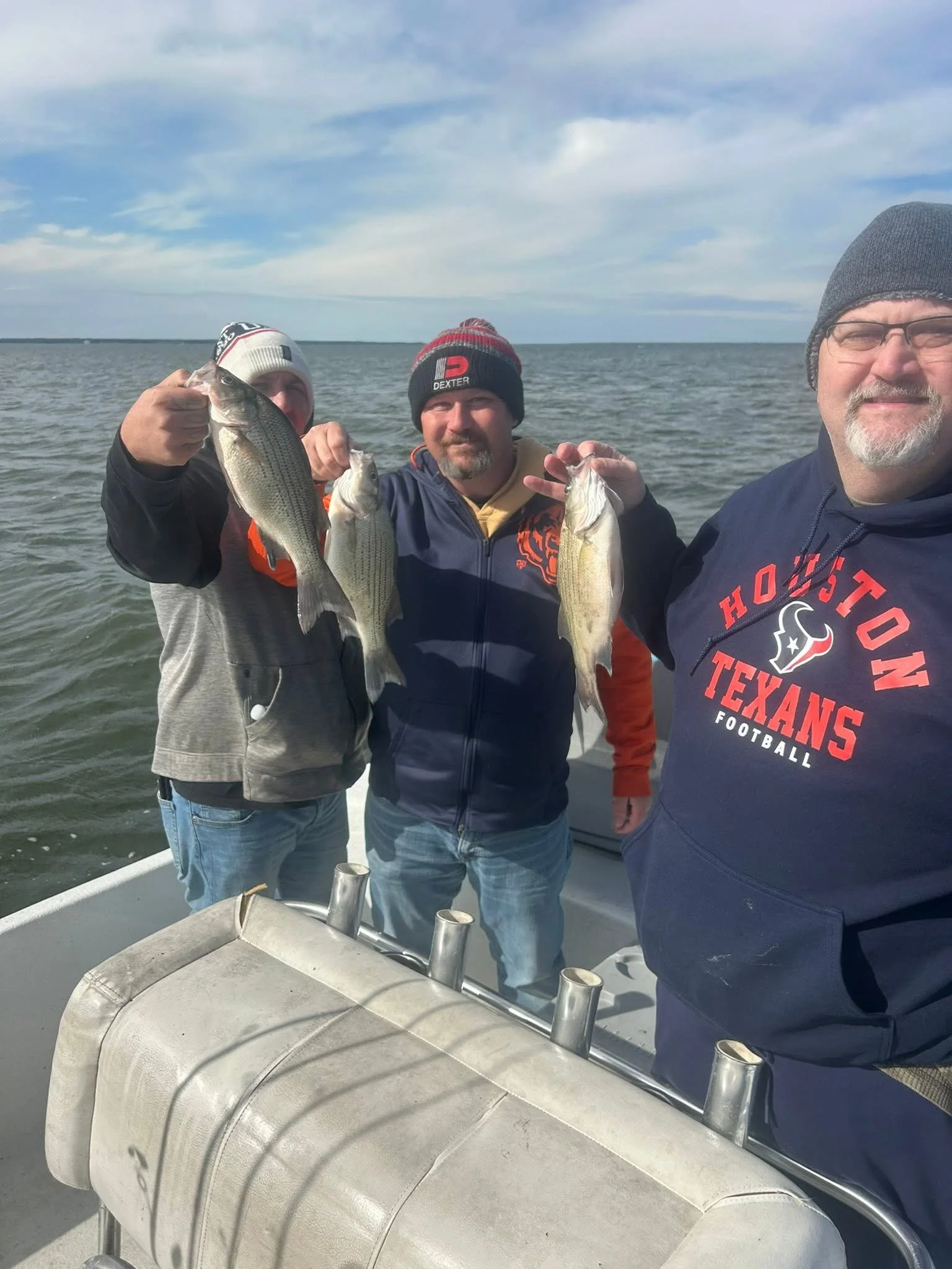 Three men standing on a boat holding up fish they caught, with a large body of water and cloudy sky behind them.