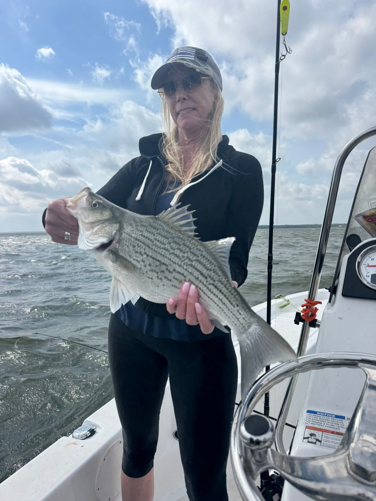 Woman on a boat holding a fish she caught, with fishing gear and water in the background.