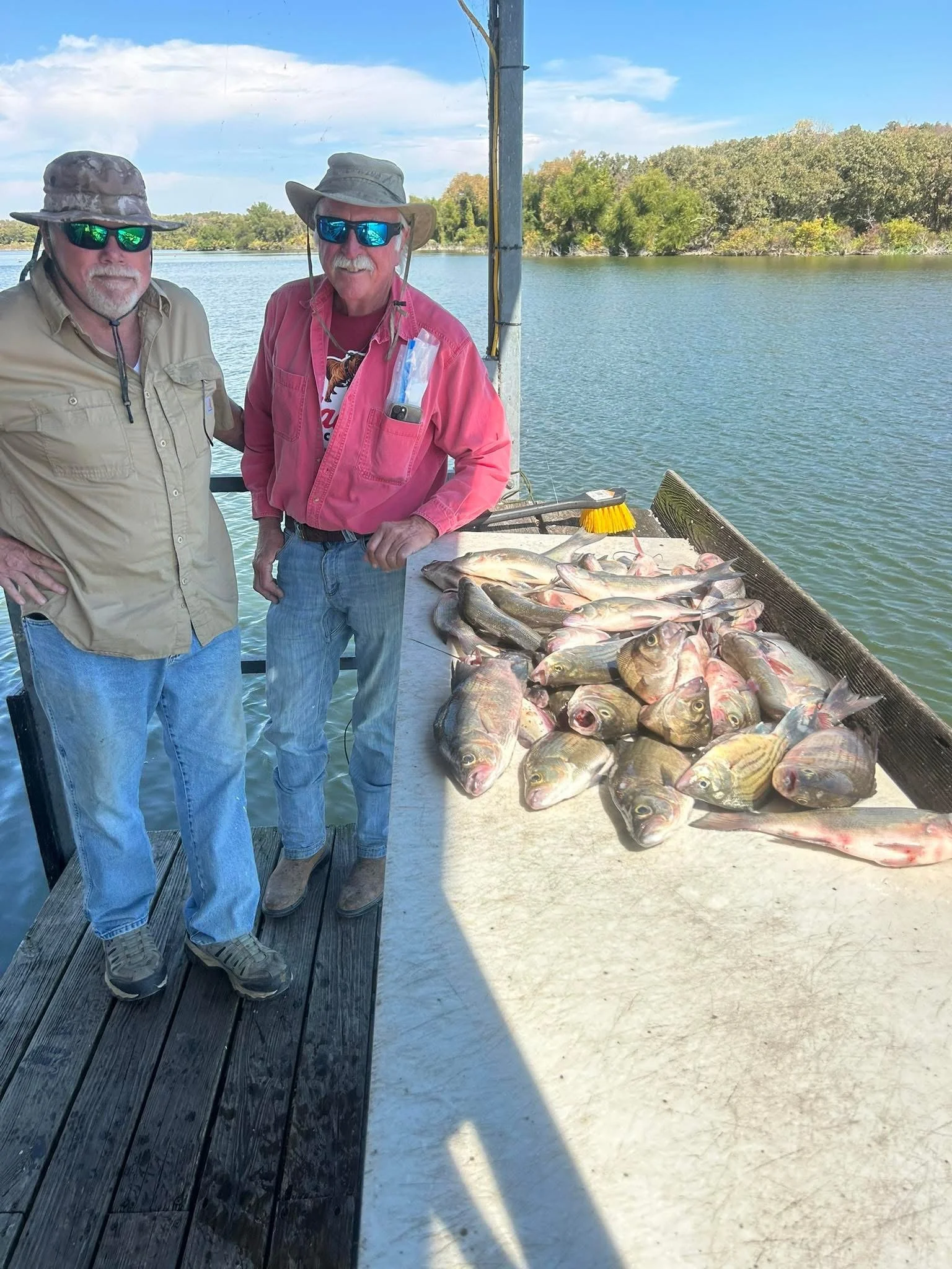 Two men wearing hats and sunglasses on a dock next to a large catch of fish laid out on a table, with water and trees in the background.
