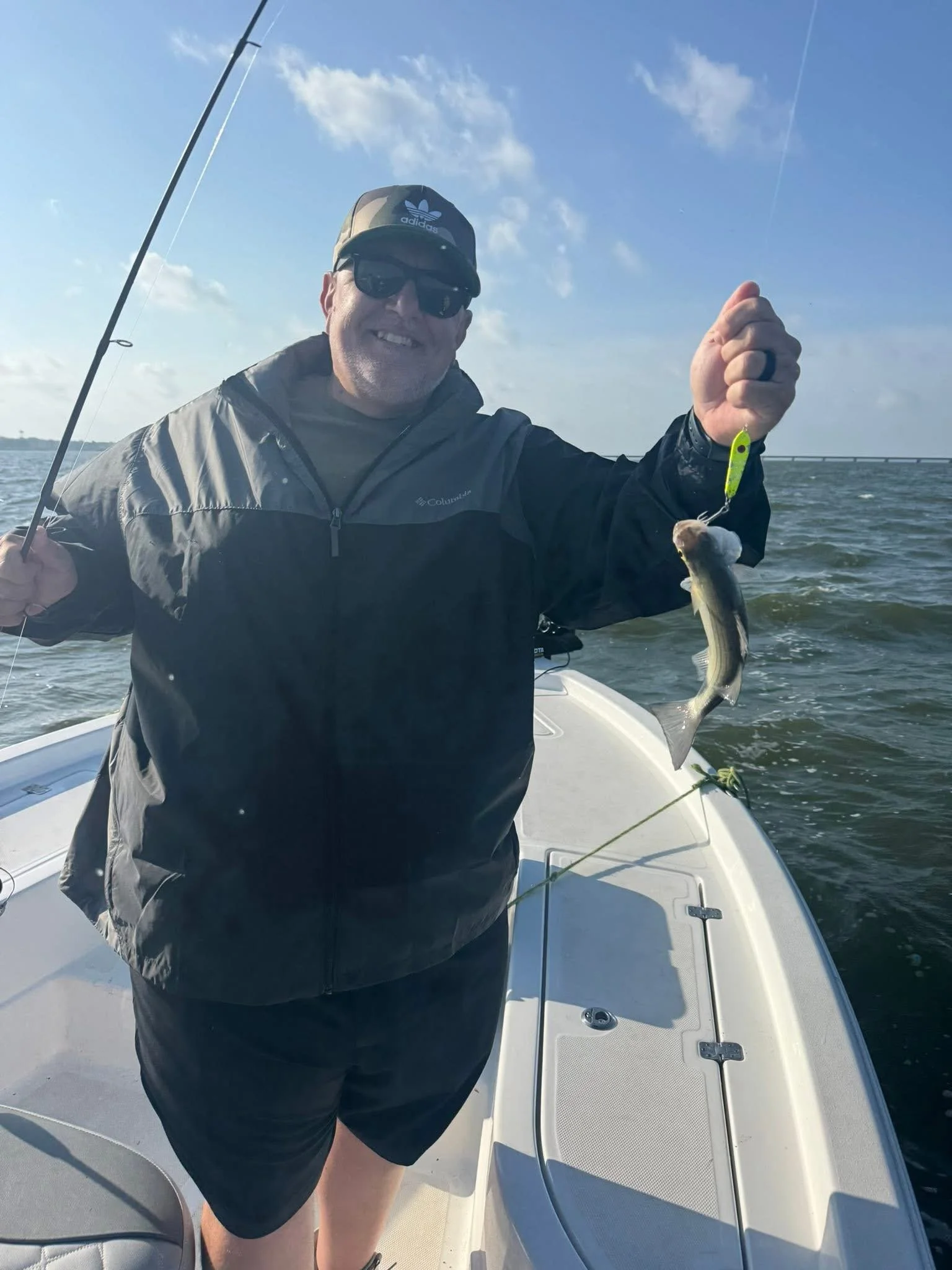 A man on a boat holding a fishing rod in one hand and a fish he caught in the other, smiling at the camera, under blue skies with clouds and a bridge in the background.