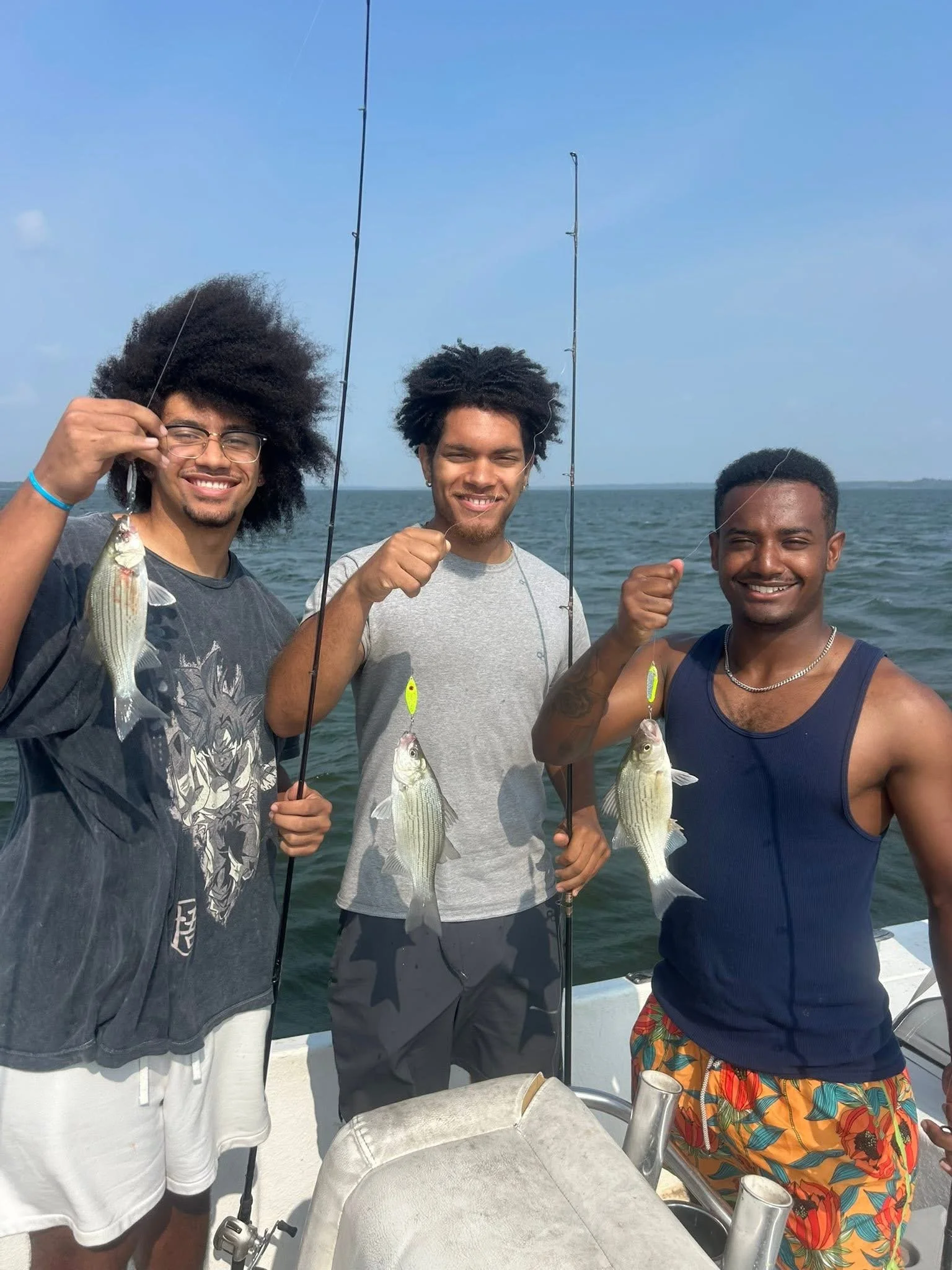 Three men on a boat holding fishing rods with fish they've caught, smiling with the ocean in the background under a partly cloudy sky.