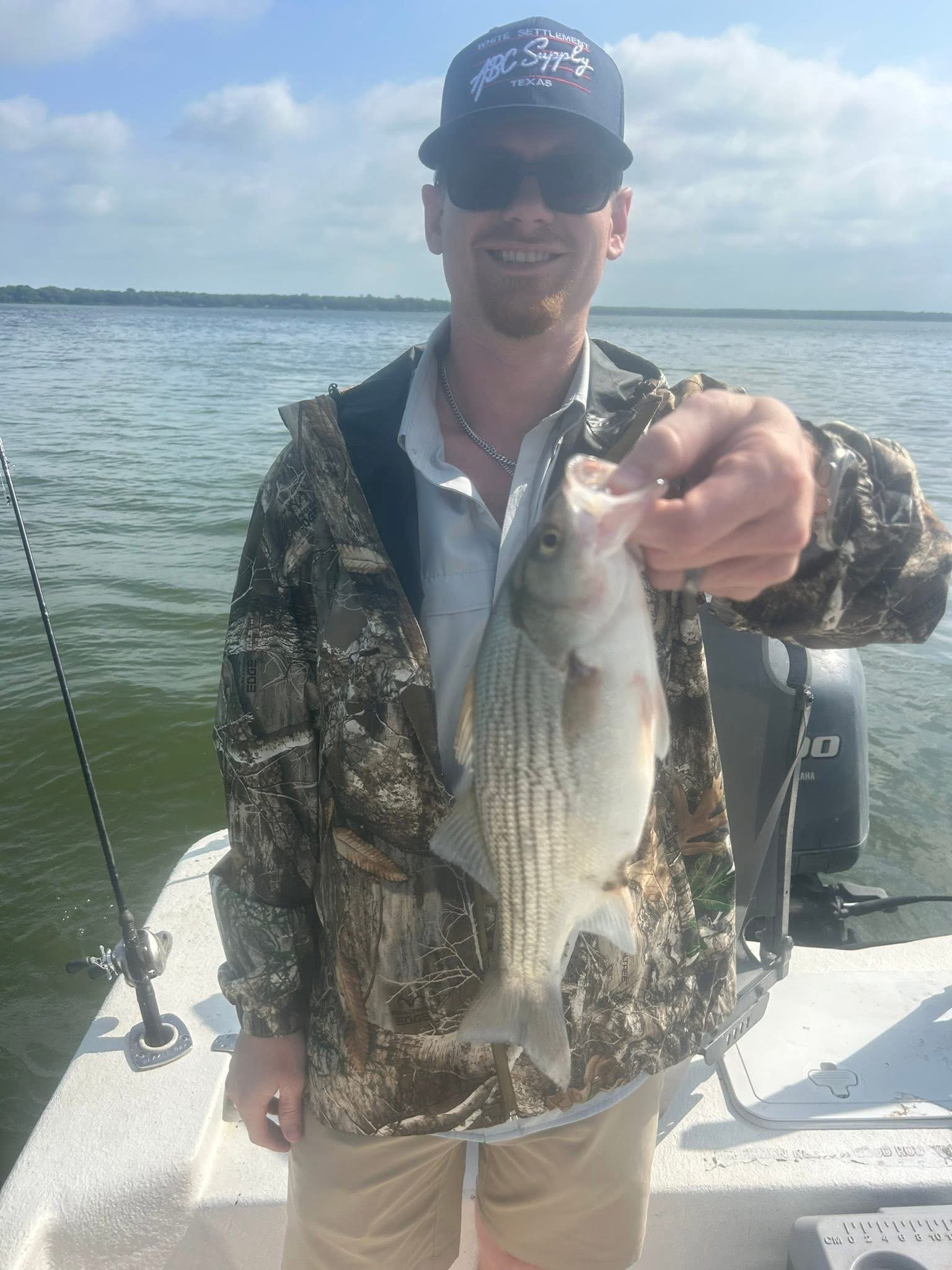 A man in camouflage jacket and beige shorts on a boat, holding a fish in front of him. He is wearing sunglasses and a black baseball cap with white and red text. The background shows a calm body of water and a partly cloudy sky.