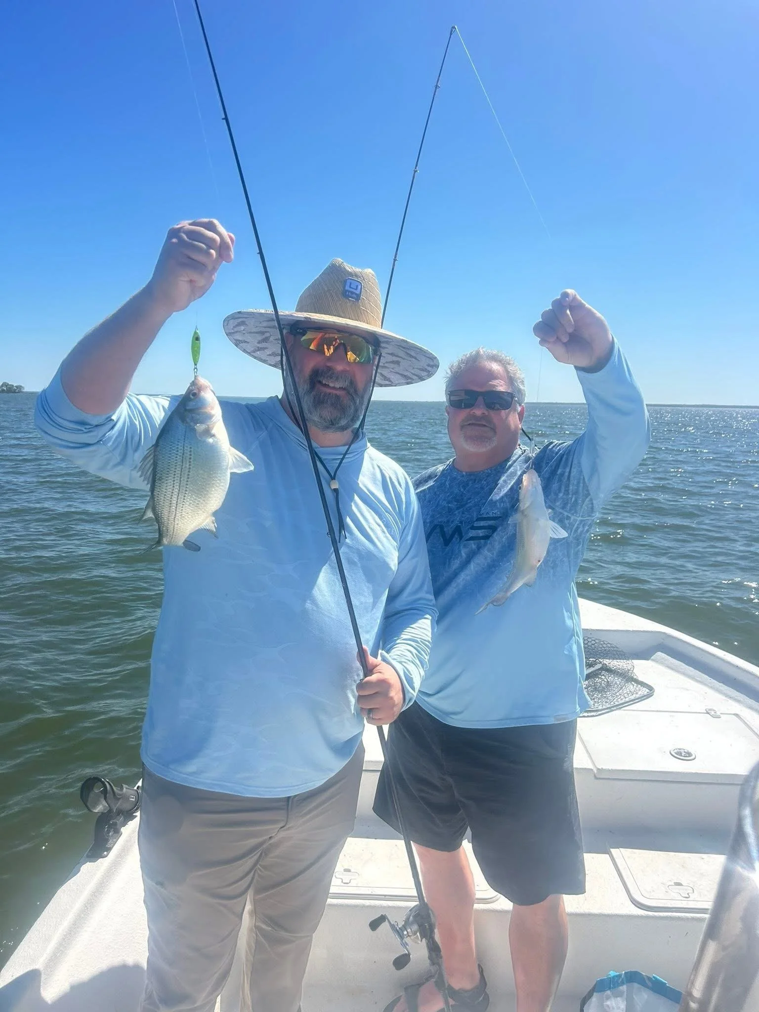 Two men on a boat holding fishing poles and a fish, with water and a clear blue sky in the background.