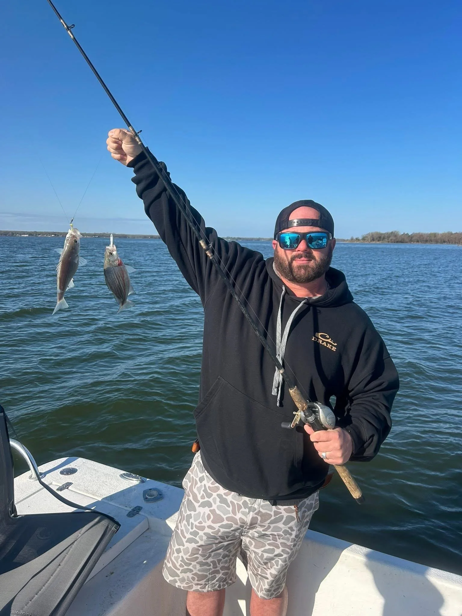 Man in a black hoodie, patterned shorts, and sunglasses holding a fishing rod with two fish caught, standing on a boat in a large body of water under a clear blue sky.