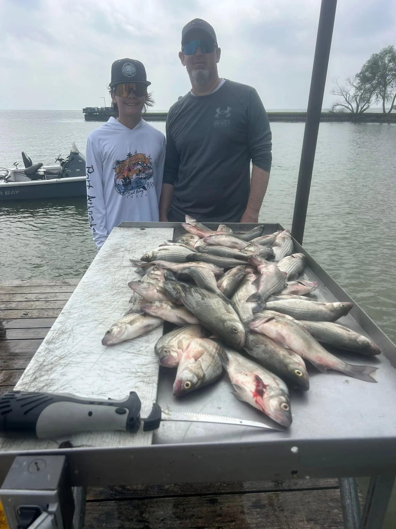 Two people standing behind a table full of freshly caught fish, near a body of water with a boat and trees in the background.
