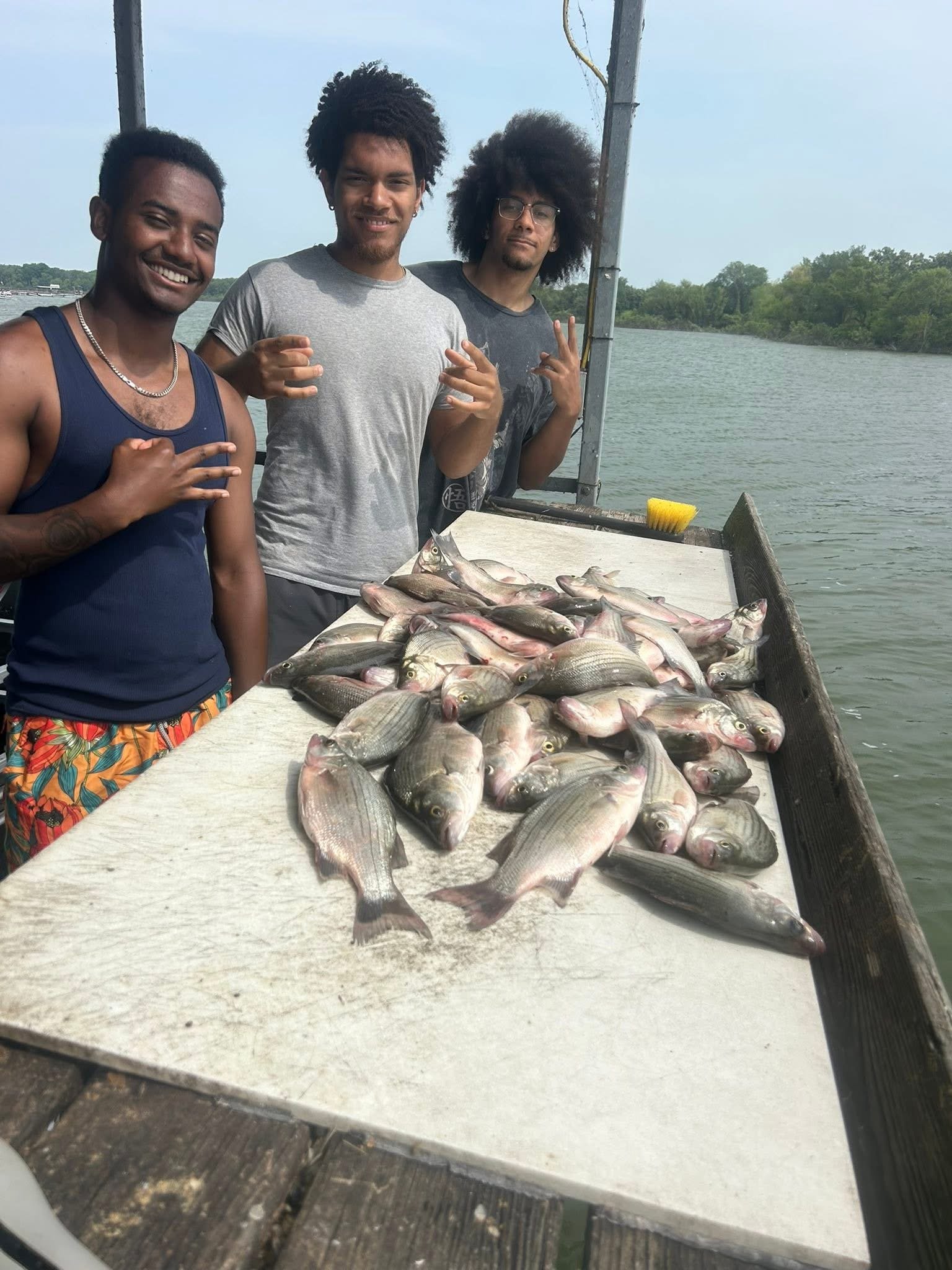 Three men standing behind a table full of freshly caught fish by a body of water, with trees in the background.