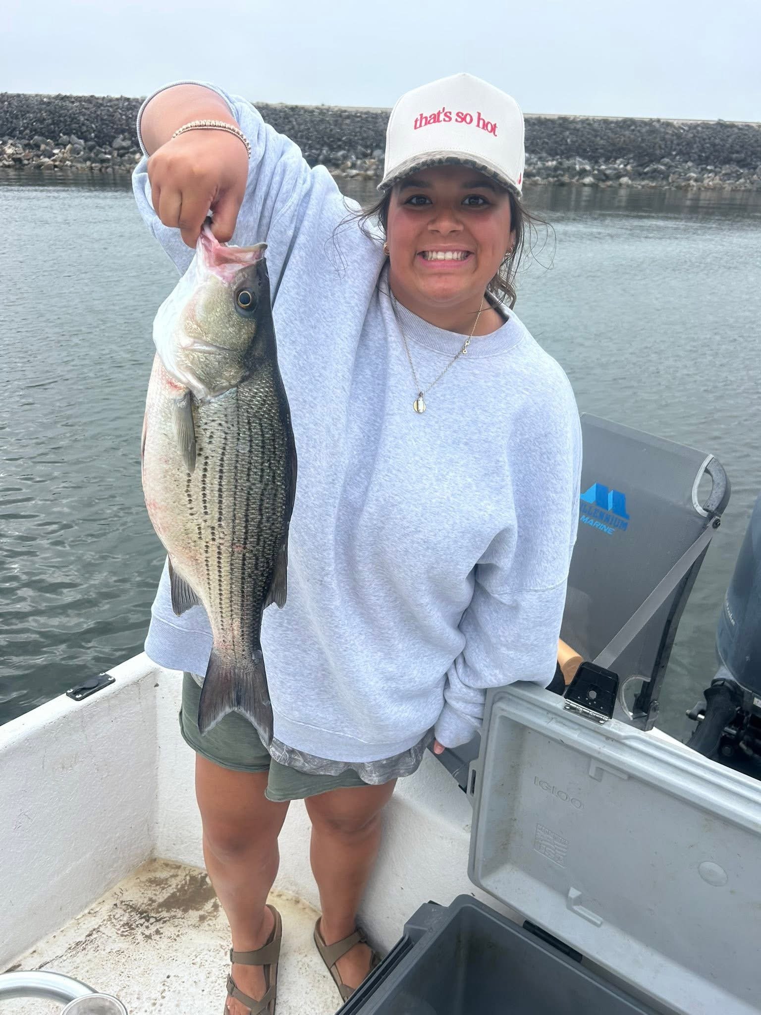 A woman smiling and holding a large fish she caught while fishing on a boat. She is wearing a white hoodie, shorts, sandals, and a cap that says 'that's so hot.' There is water and a rocky shoreline in the background.