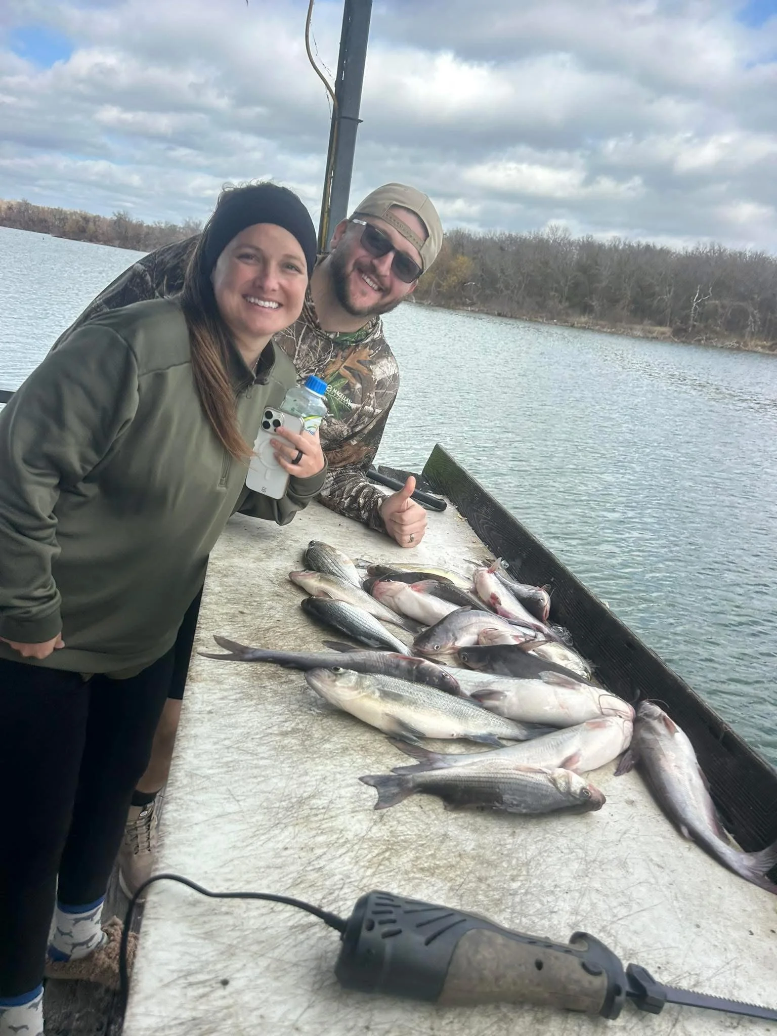 Two people smiling near a collection of caught fish on a dock by a body of water, with cloudy skies overhead.